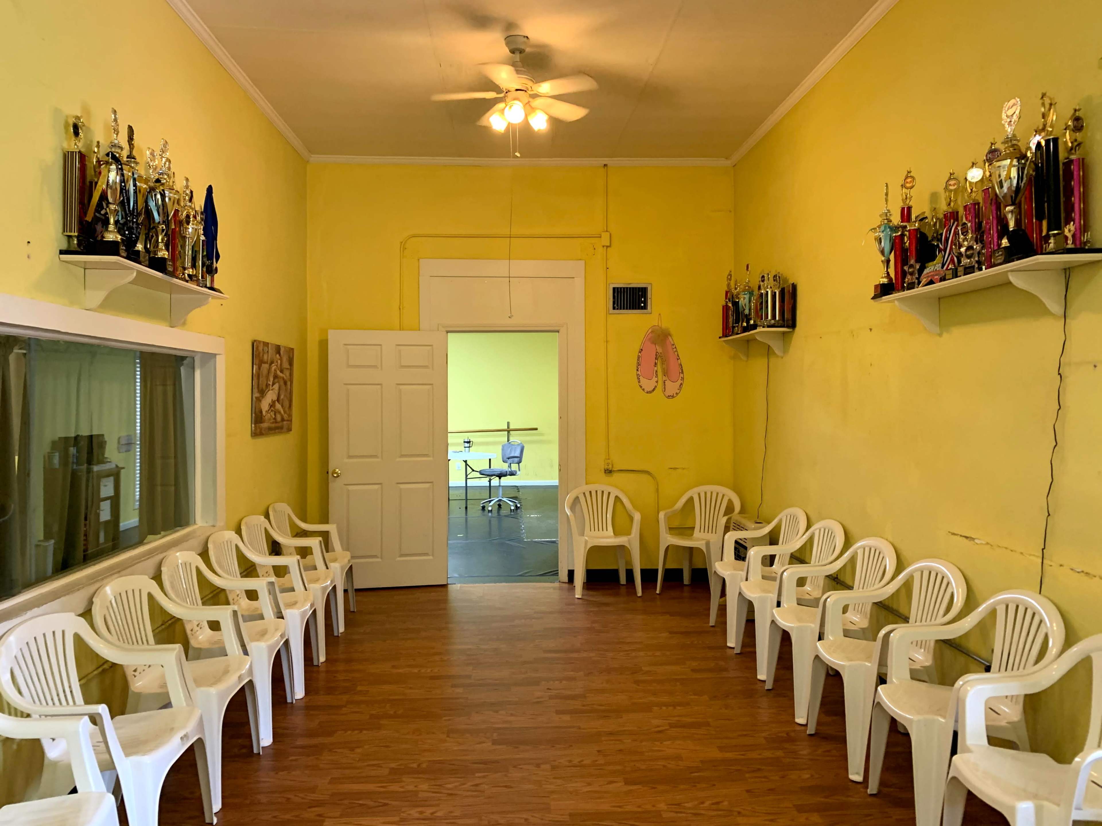 A room with yellow walls features two rows of white plastic chairs and shelves displaying various trophies.
