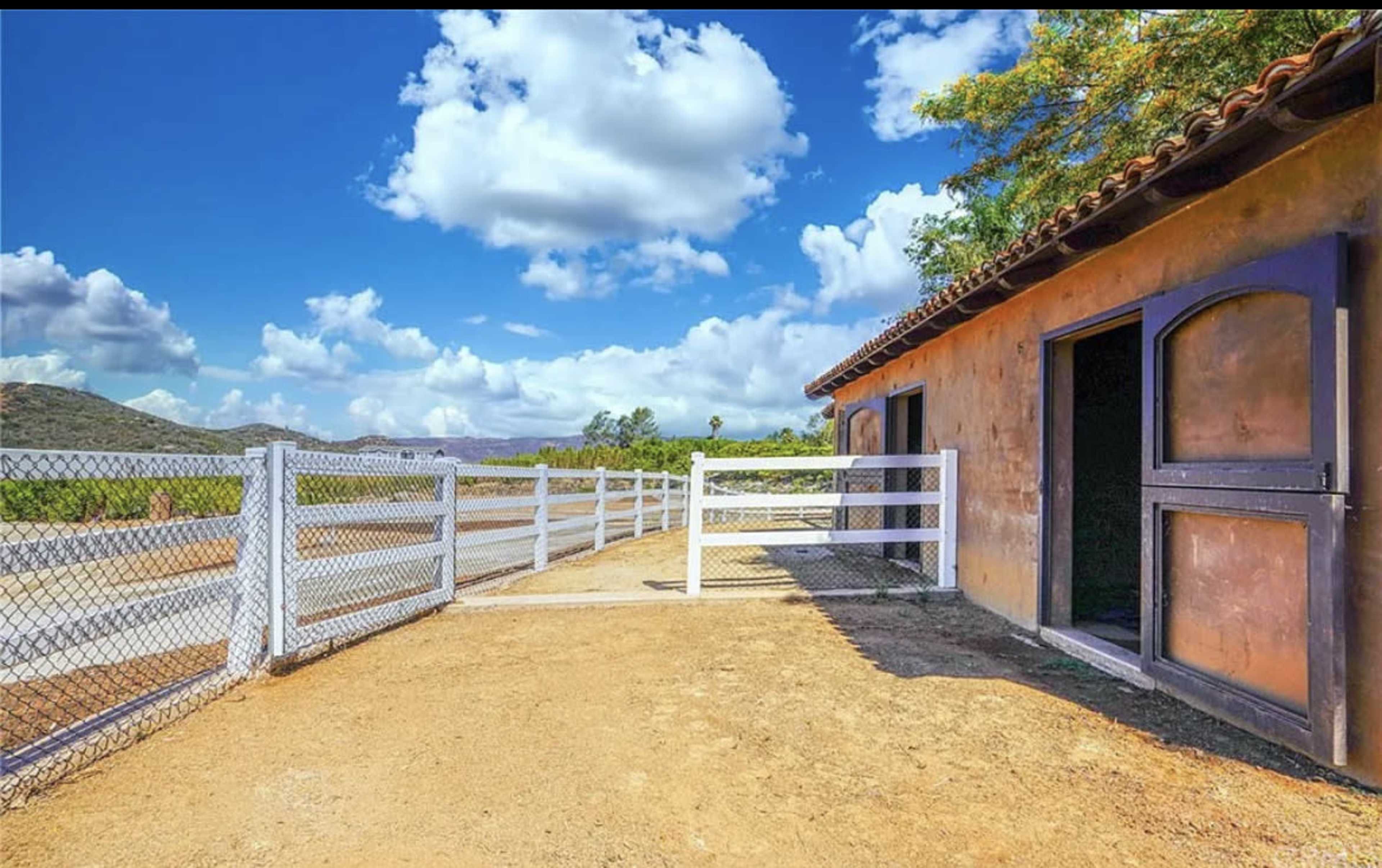 A rustic building with open doors is set beside a white fence under a blue sky with fluffy clouds.