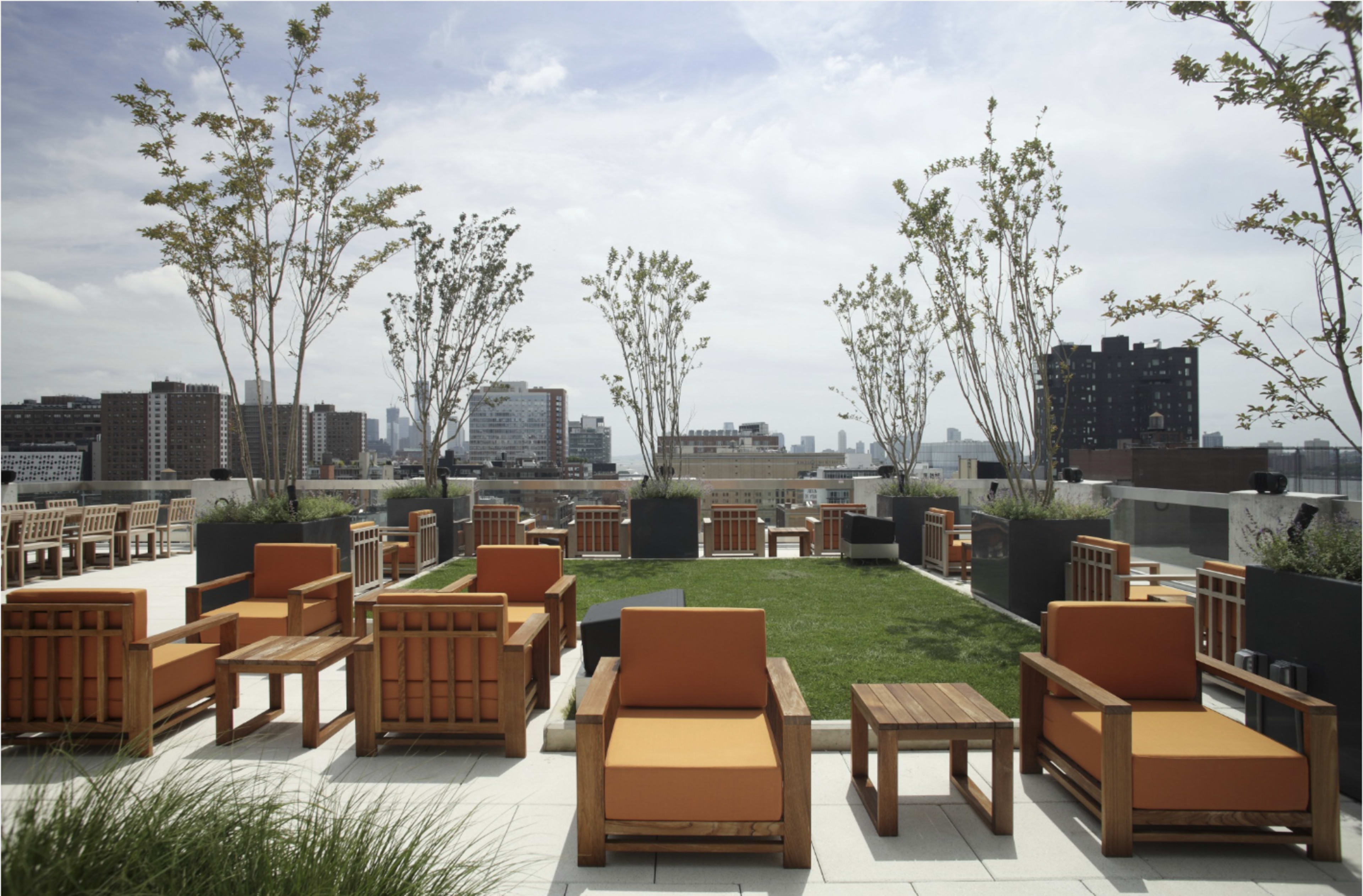 The image shows a rooftop terrace with wooden lounge chairs and tables arranged on a deck beside manicured grass areas and several trees.