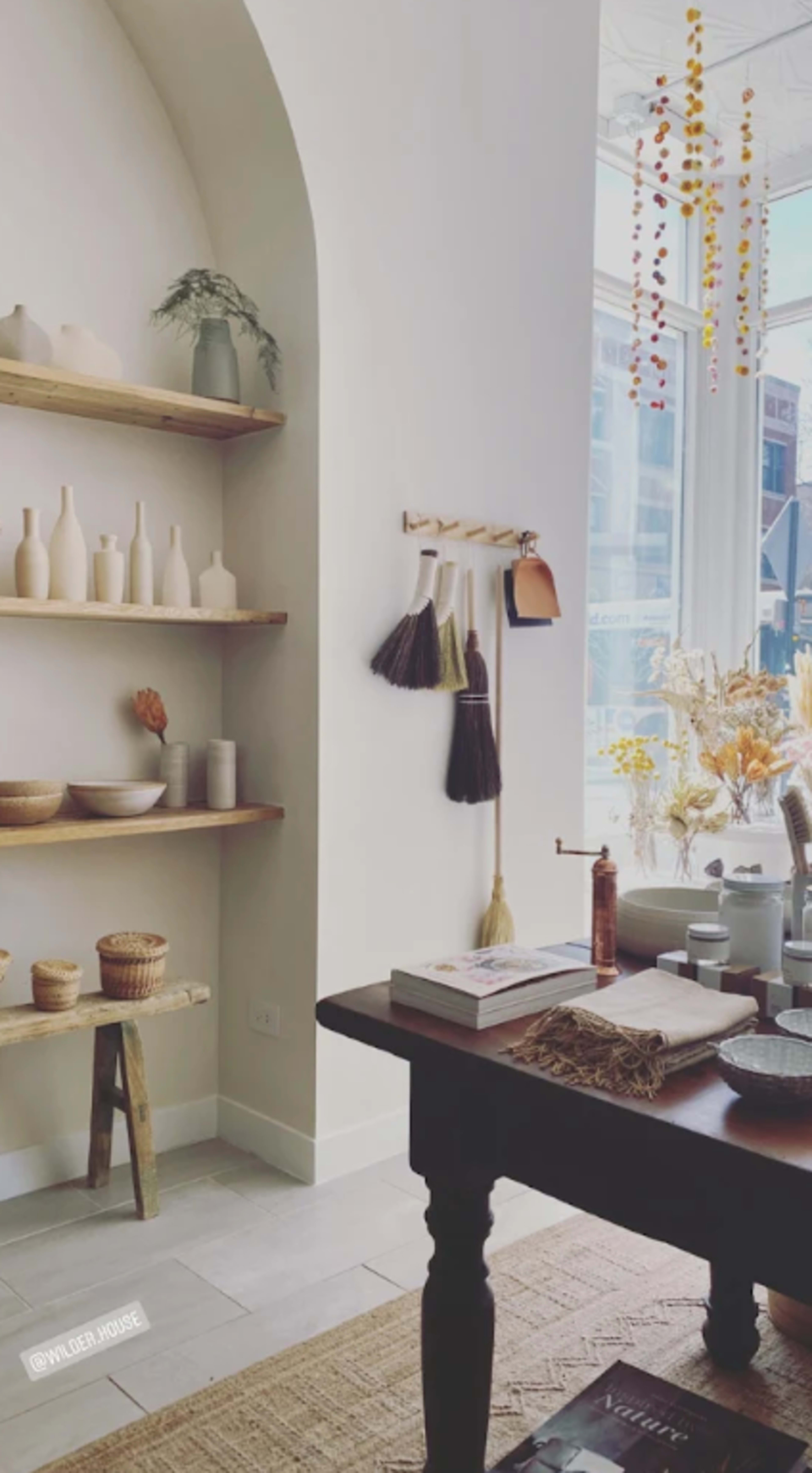 The image shows a light-filled interior featuring wooden shelves with decorative vases, a table with various pottery items, and hanging dried flowers near a large window.
