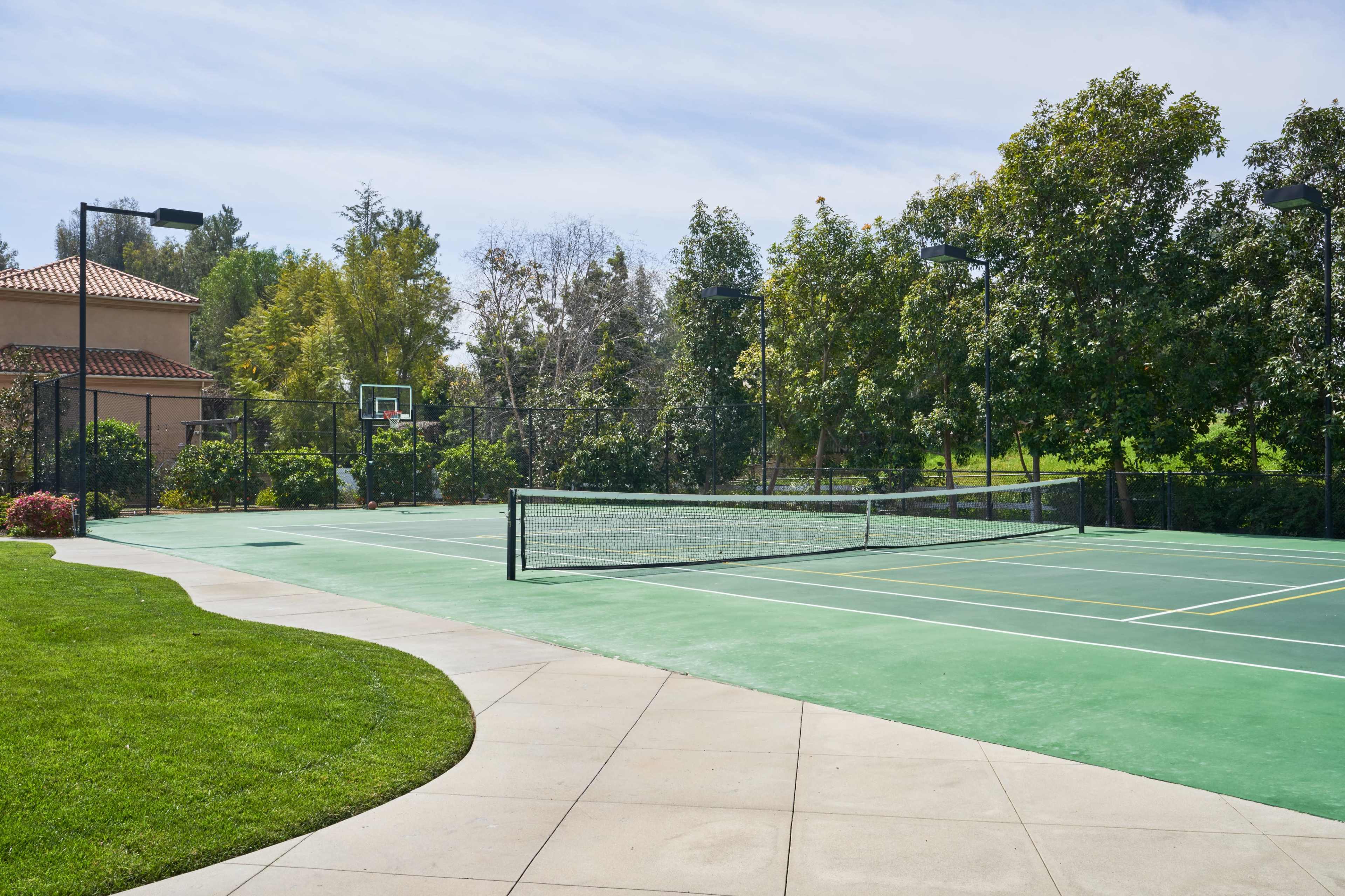 A well-maintained tennis court beside a basketball hoop, surrounded by greenery and pathways.