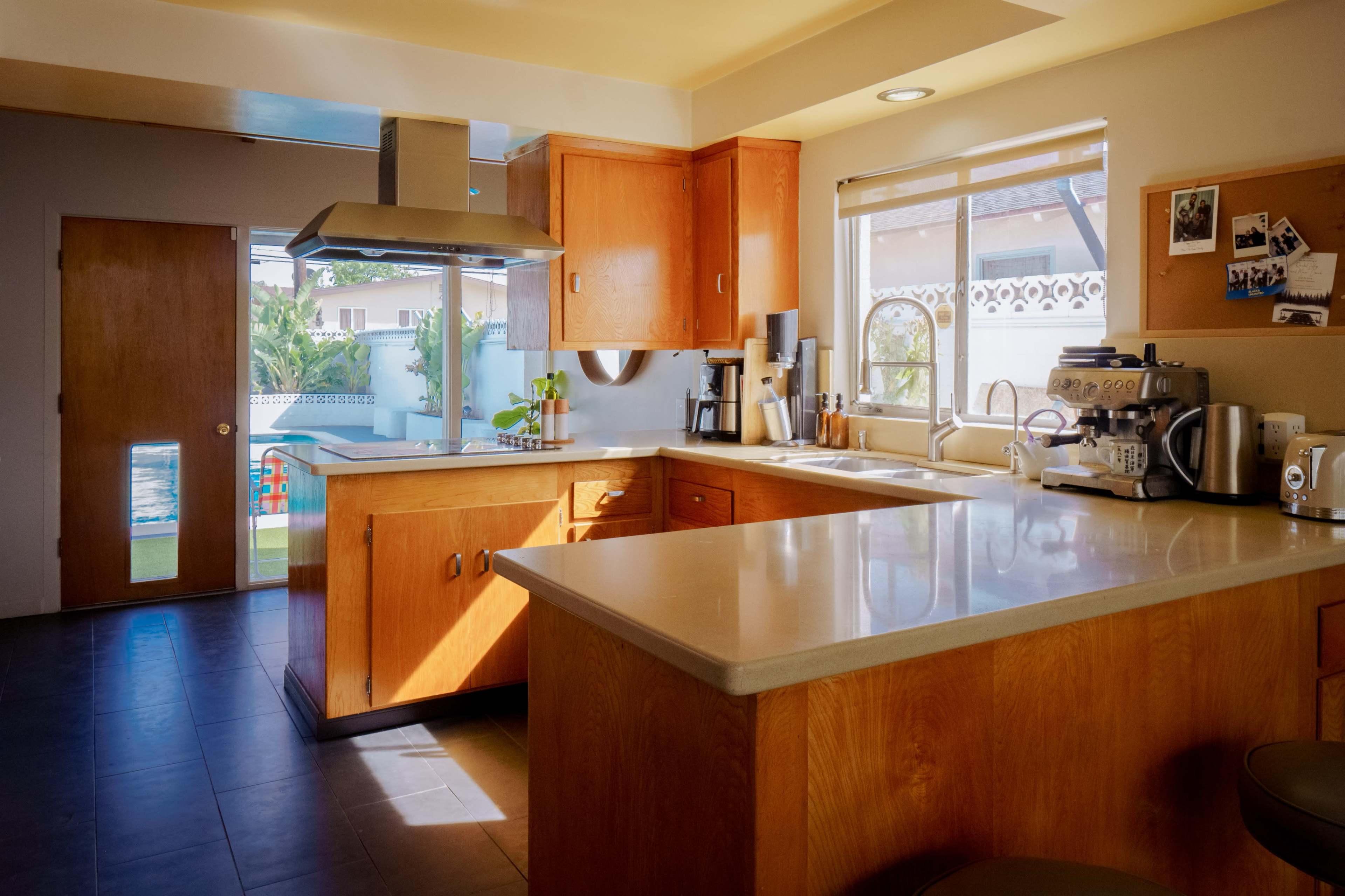 A modern kitchen with wooden cabinets, a stainless steel range hood, and a countertop that features a coffee machine and various kitchen tools, illuminated by natural light.