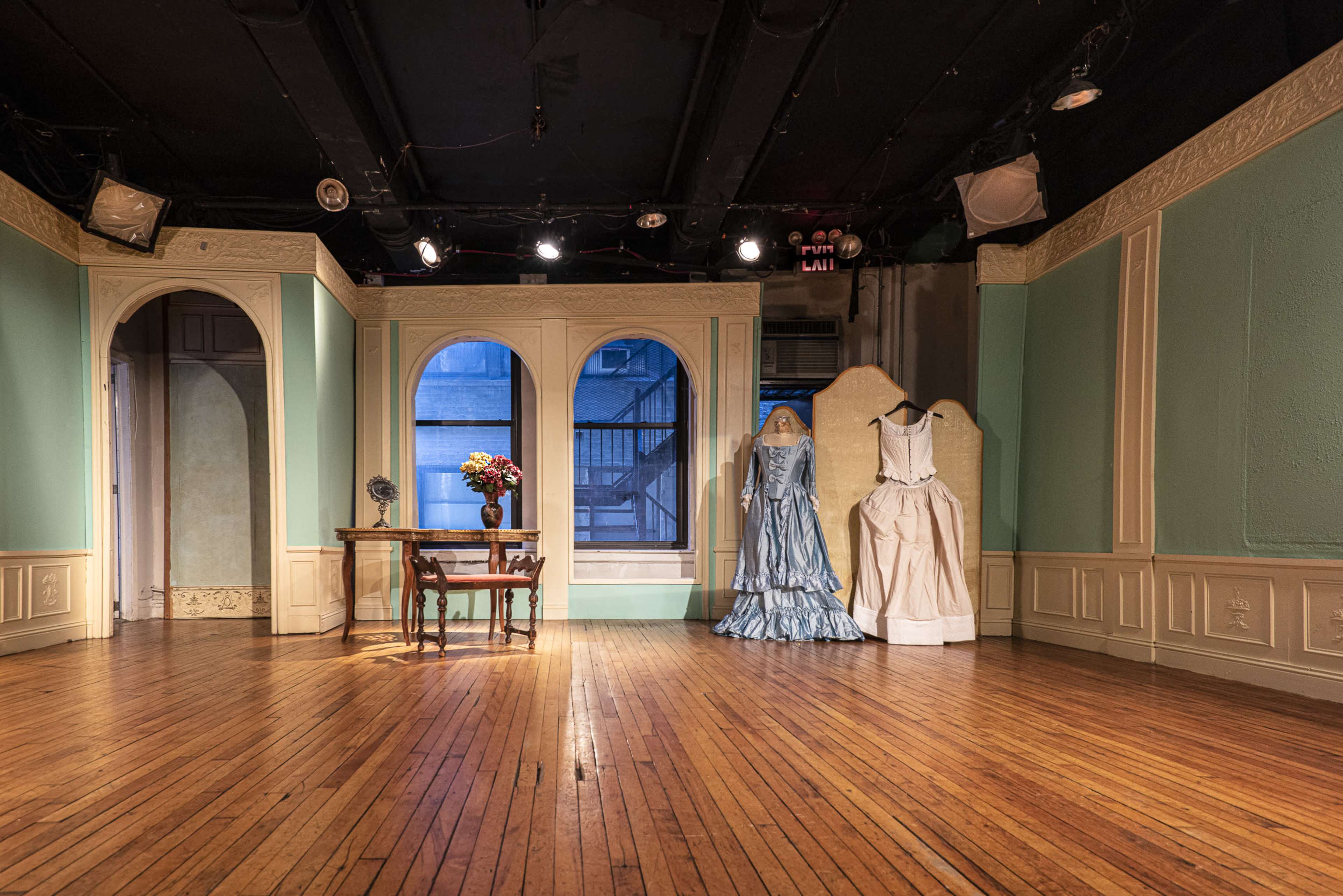 The image shows a stage setting with a wooden floor, two elegant dresses displayed on mannequins, a small table with flowers, and arched windows revealing a view of an exterior space.