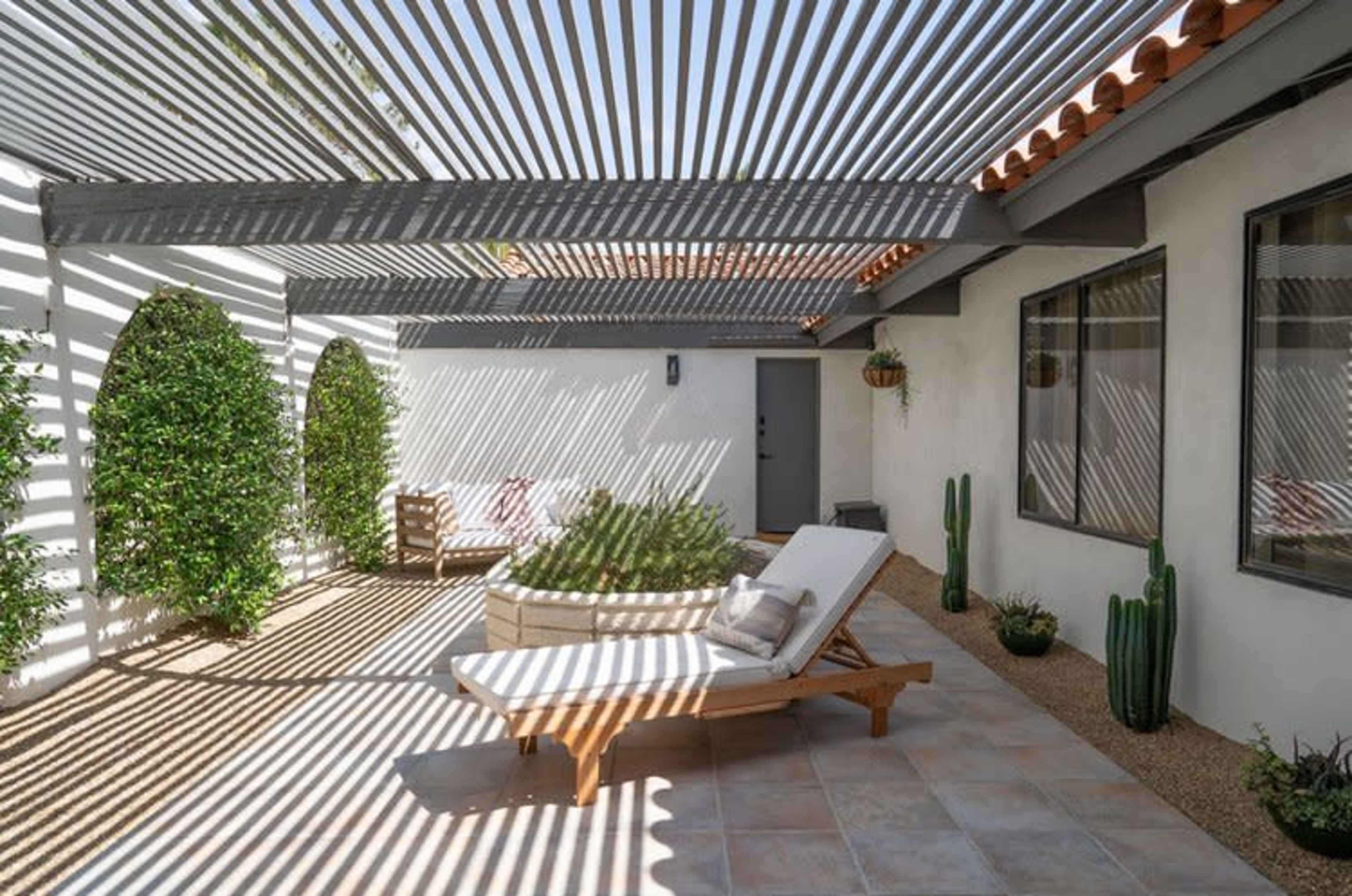 The image shows a patio with a lounge chair and a circular seating area, surrounded by potted plants and cast shadows from a slatted roof.