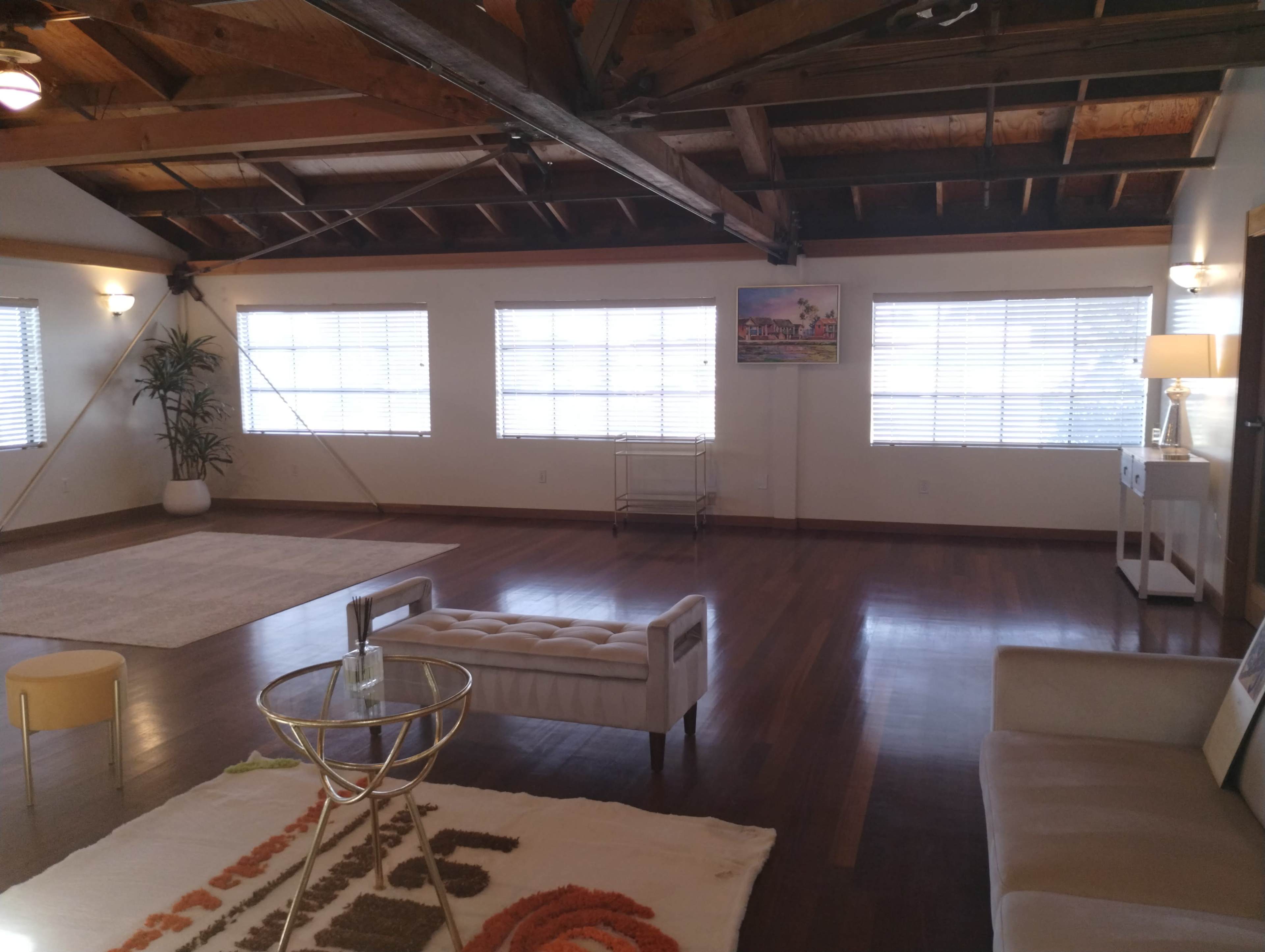 A spacious room with wooden beams, large windows covered by blinds, and minimalist furniture including a tufted sofa and a coffee table.