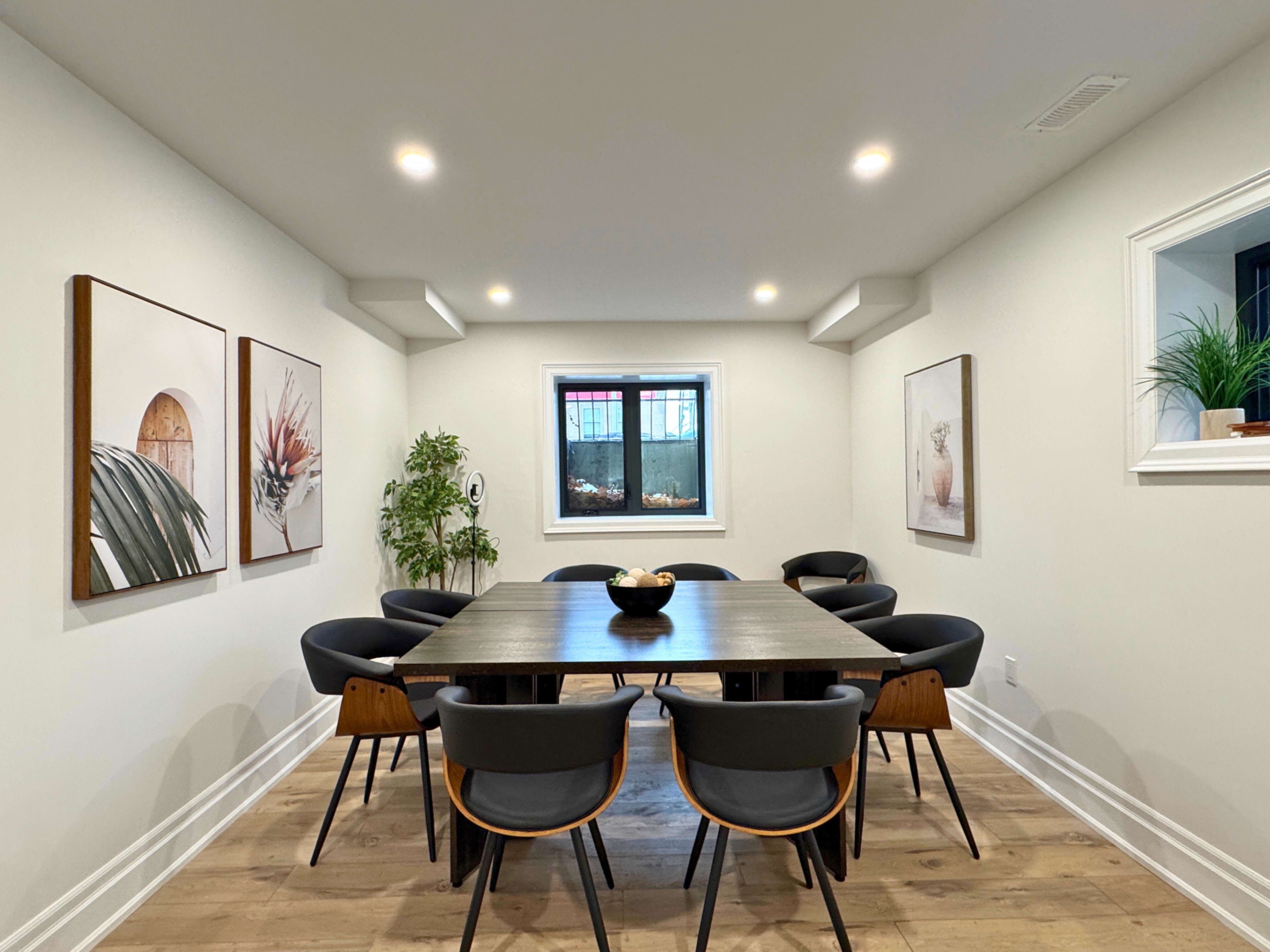 A dining room features a large table surrounded by black chairs, with framed artwork on the walls and a window providing natural light.