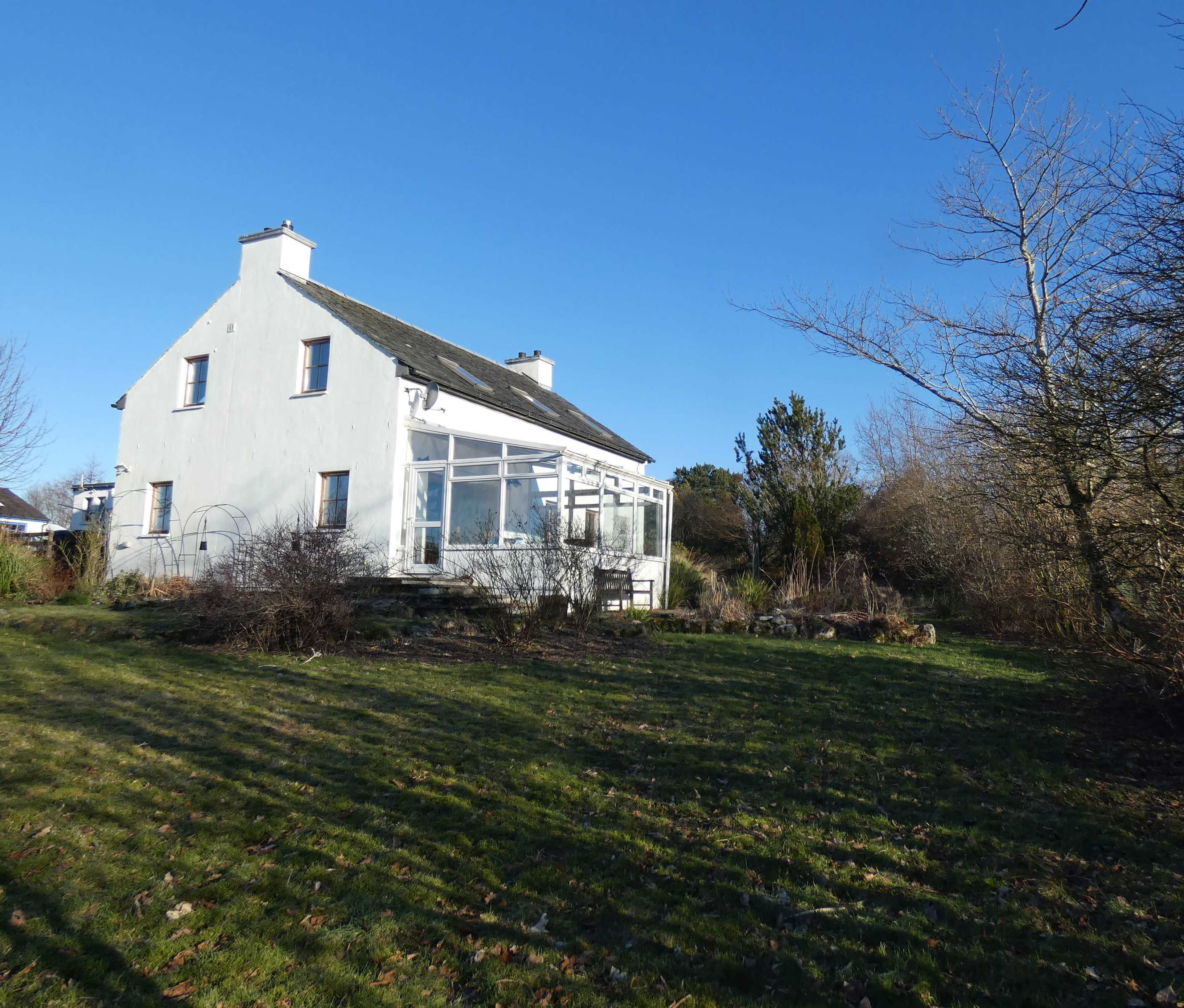 A white two-story house with a glass conservatory sits on a grassy area surrounded by sparse trees and shrubs under a clear blue sky.
