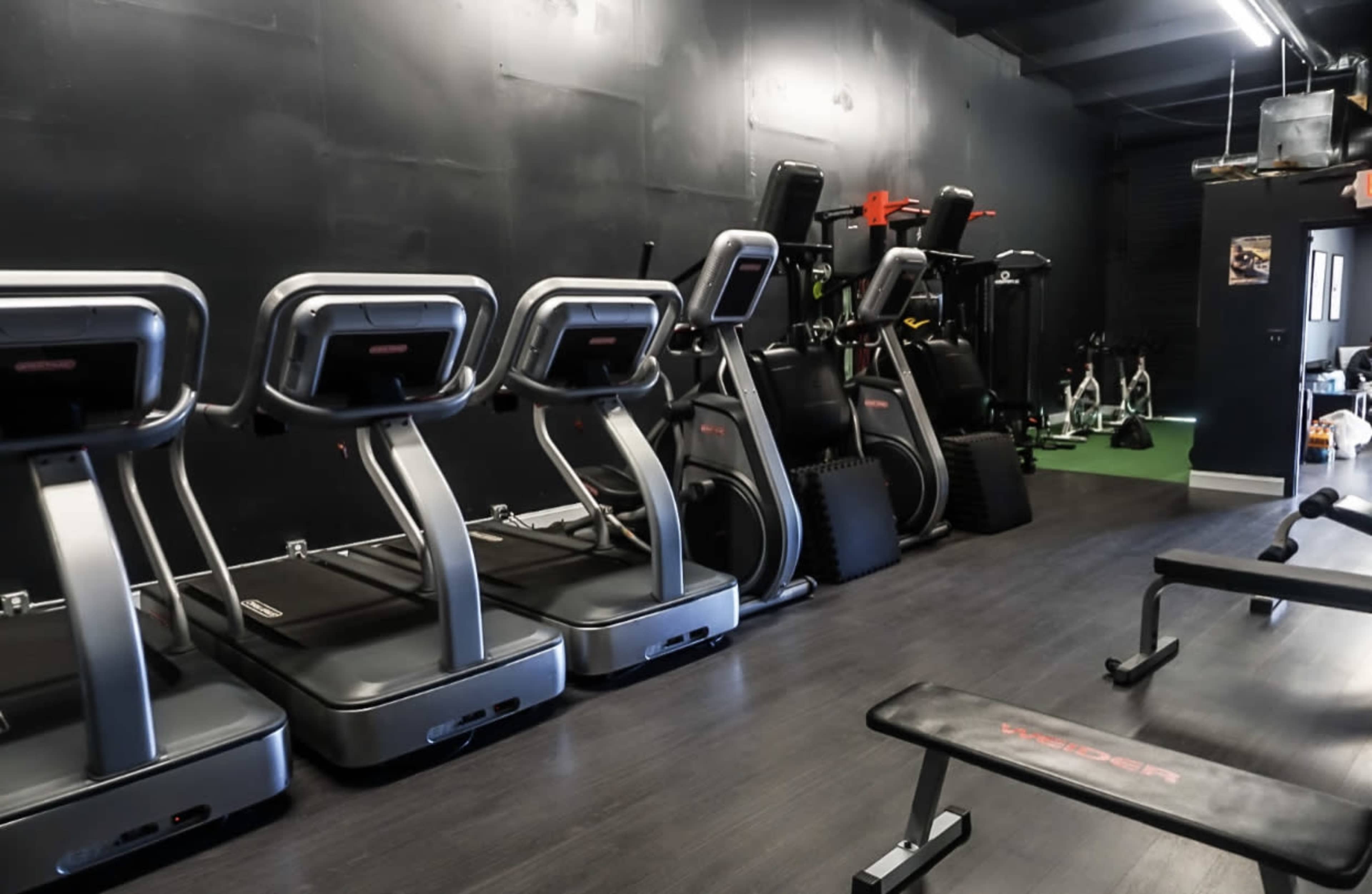 A gym interior featuring multiple treadmills lined up against a black wall, with various exercise equipment stored in the background.