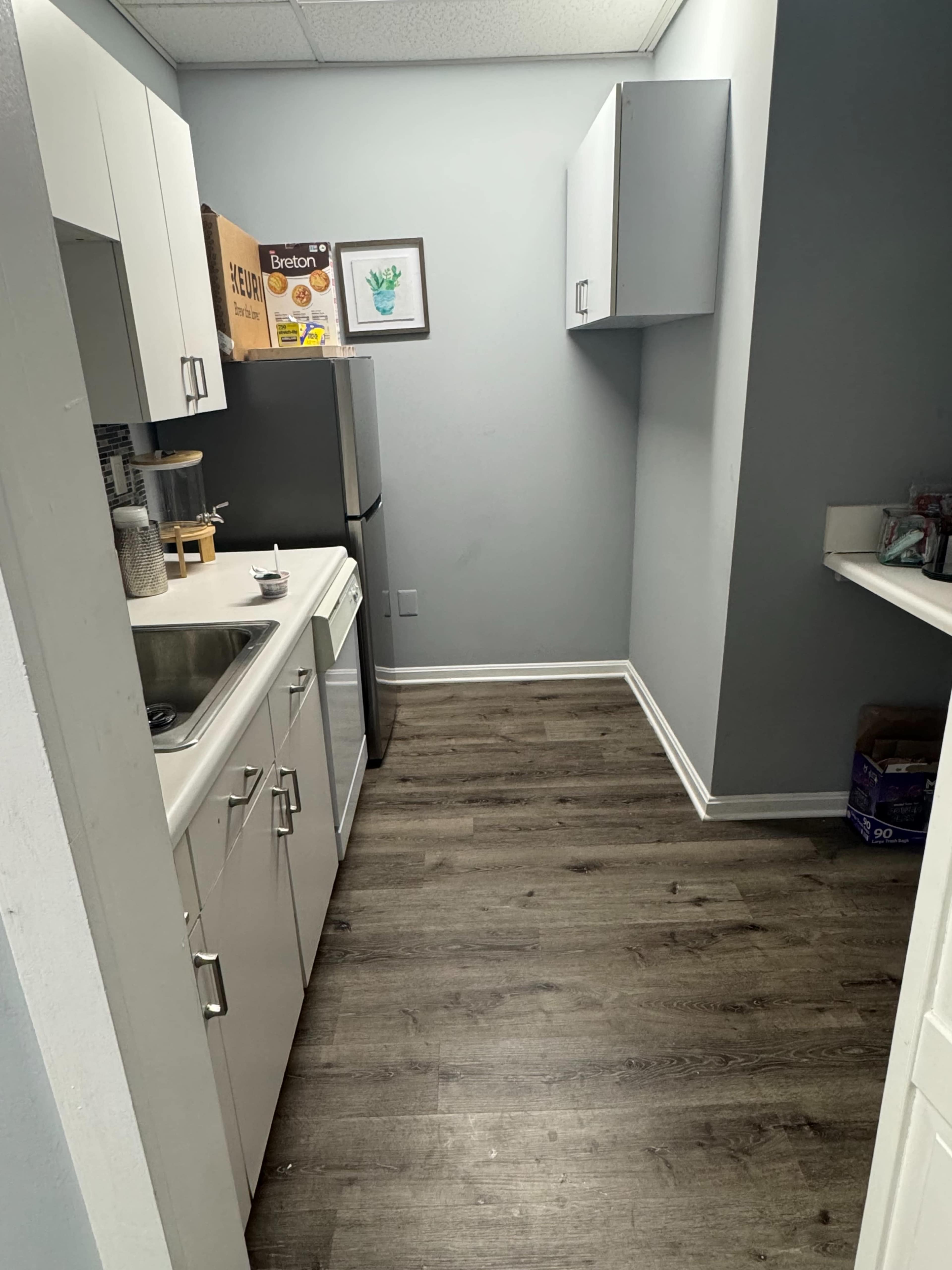 The image shows a narrow kitchen with a sink, refrigerator, and cabinets against light blue walls and a wooden floor.
