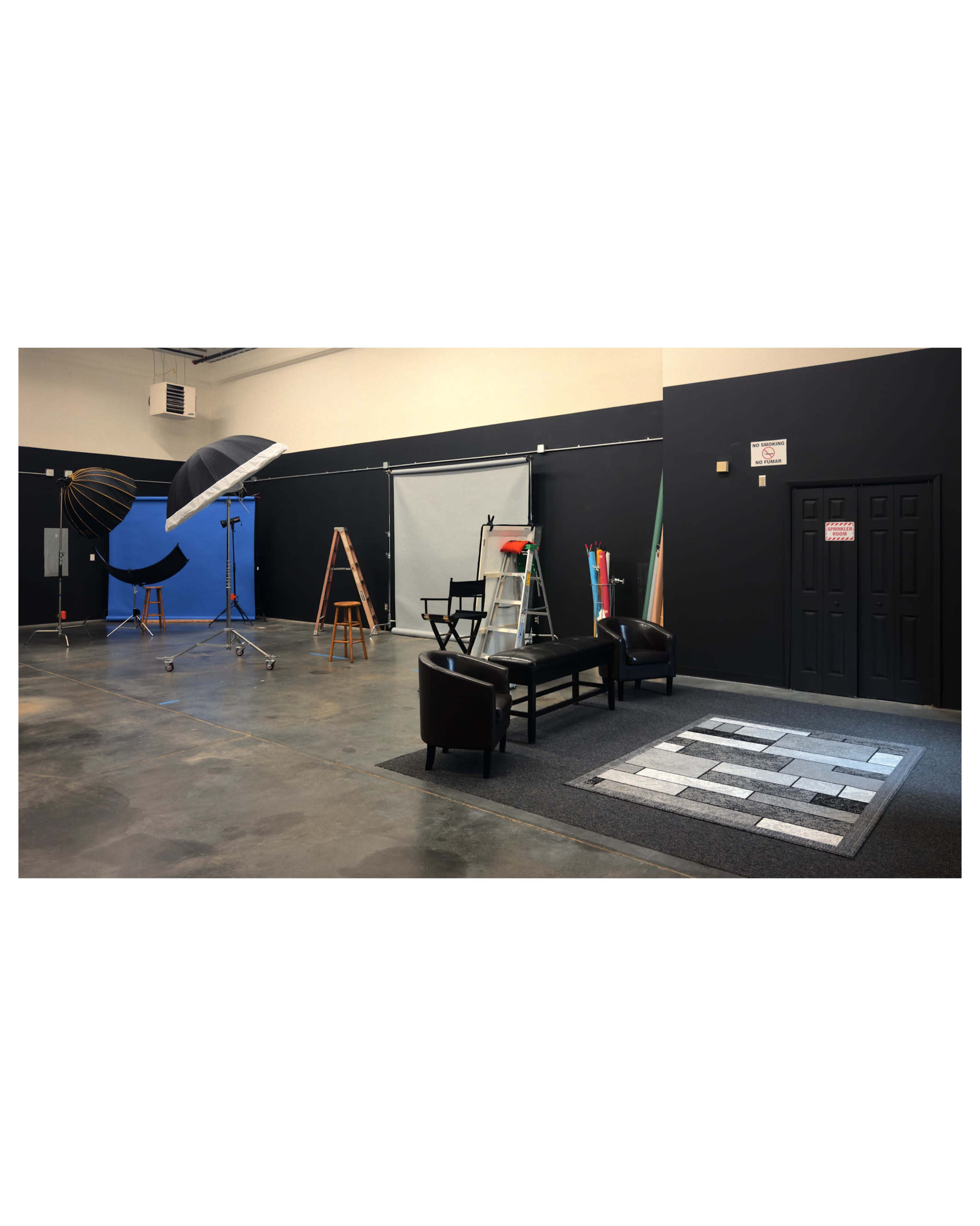 The image shows a spacious photography studio equipped with lighting equipment, a ladder, a backdrop, and a seating area with two chairs and a rug on the floor.