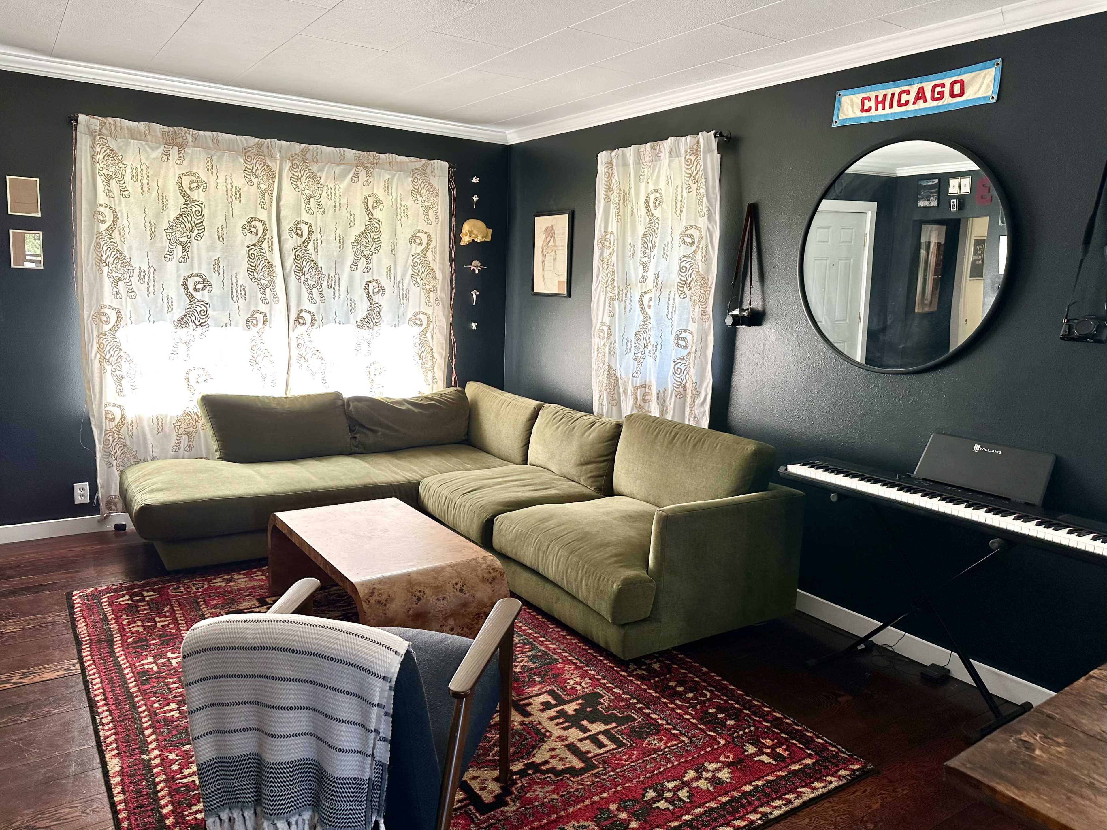 A living room with a green sectional sofa, a round mirror, a piano keyboard, and patterned curtains, all set against dark walls and a wooden floor.