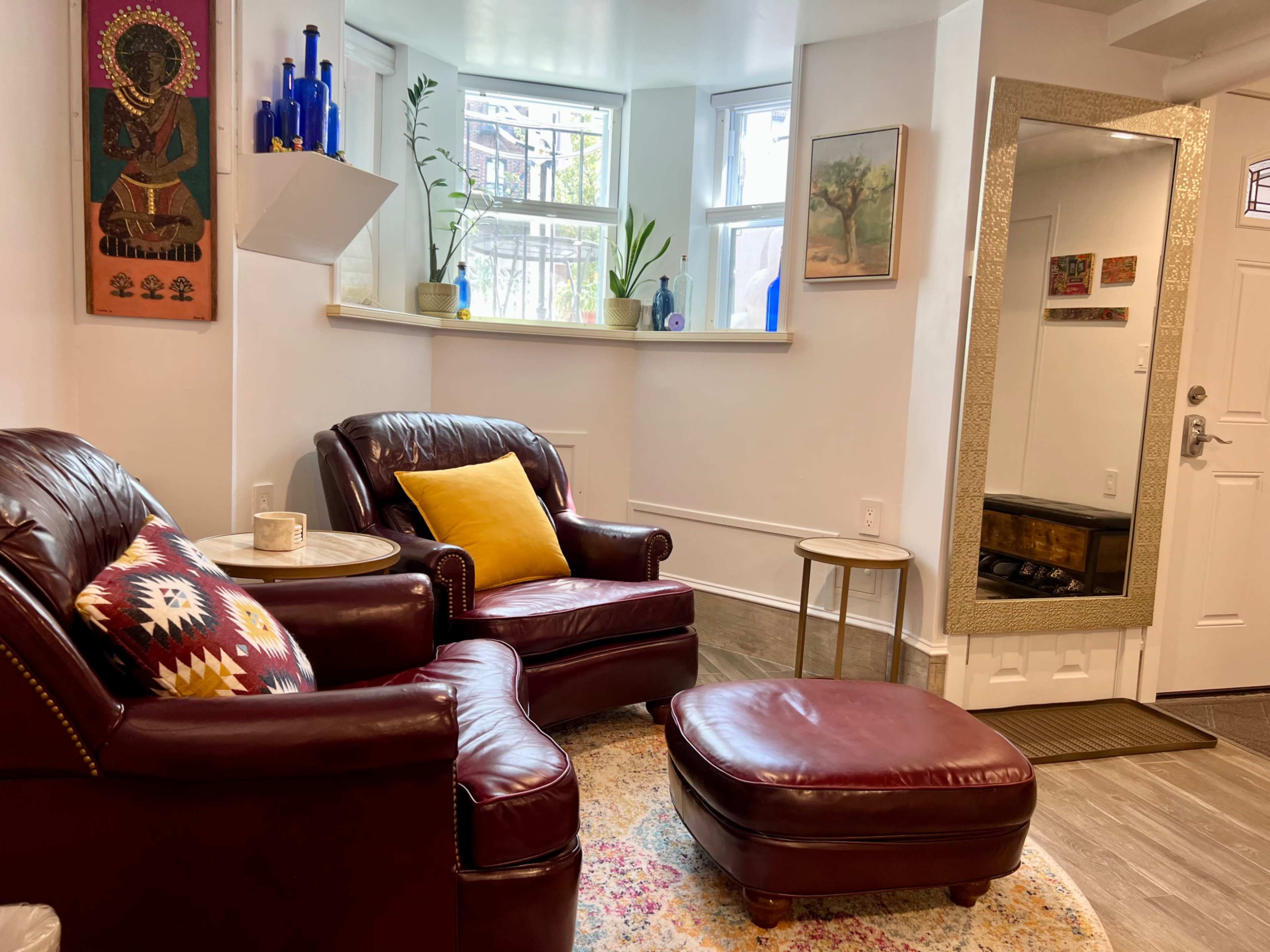 The image shows a cozy living room with two burgundy armchairs, a small round table, an ottoman, and a large mirror, surrounded by potted plants and decorative artwork.