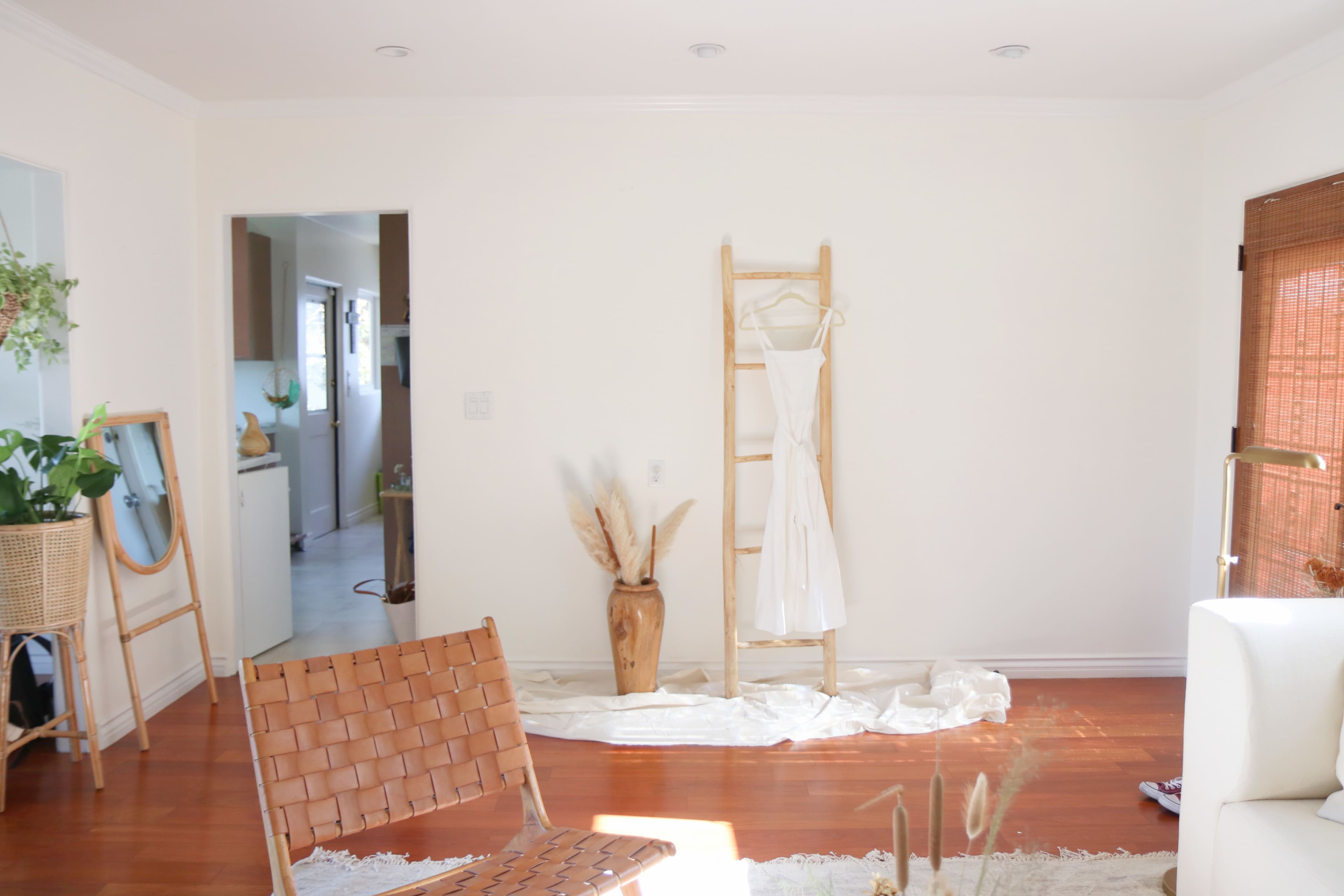 A minimalist room features a wooden ladder displaying a white dress, with a potted plant and a doorway leading to a kitchen in the background.