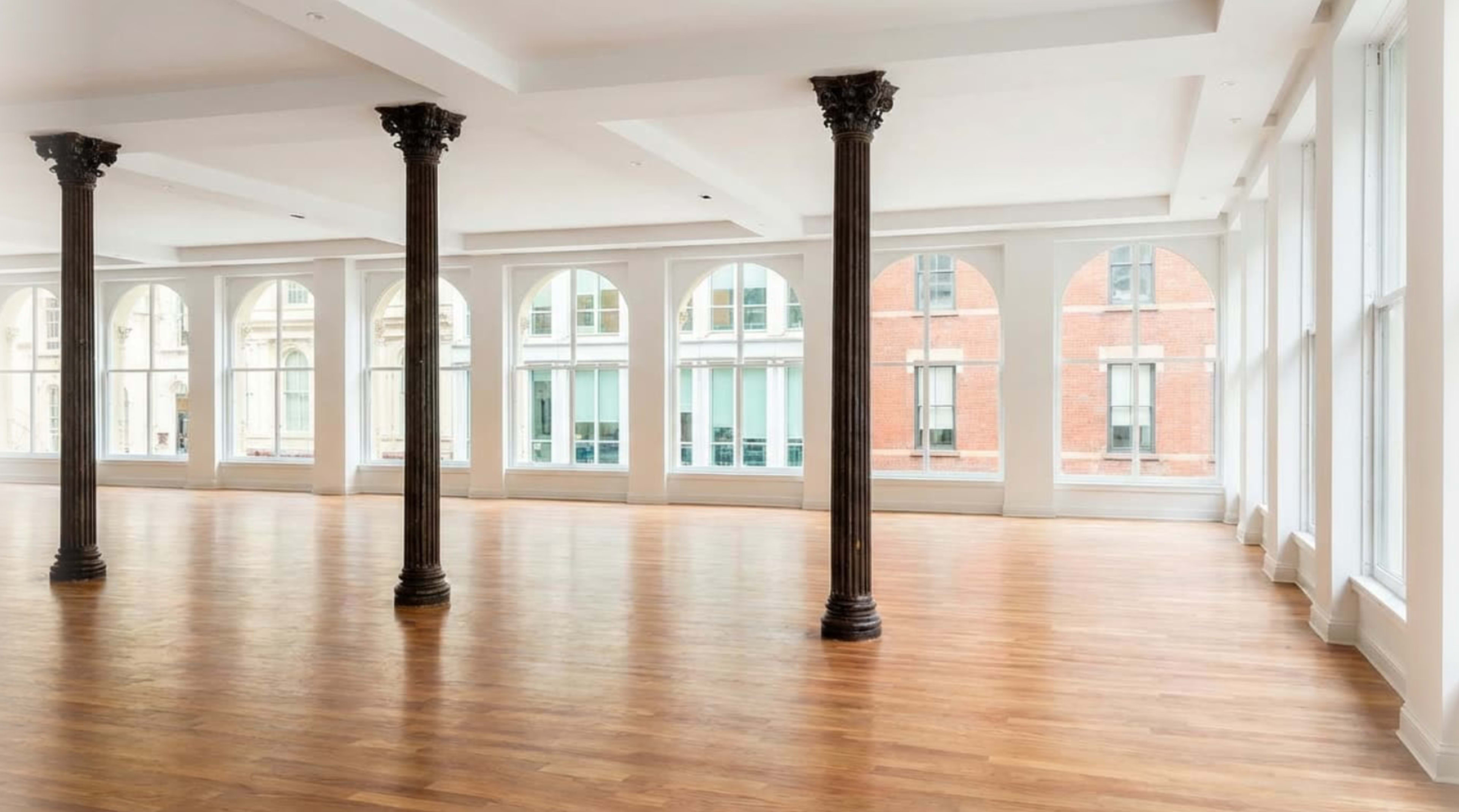 The image shows an empty interior space with wooden floors, large arched windows, and decorative columns.