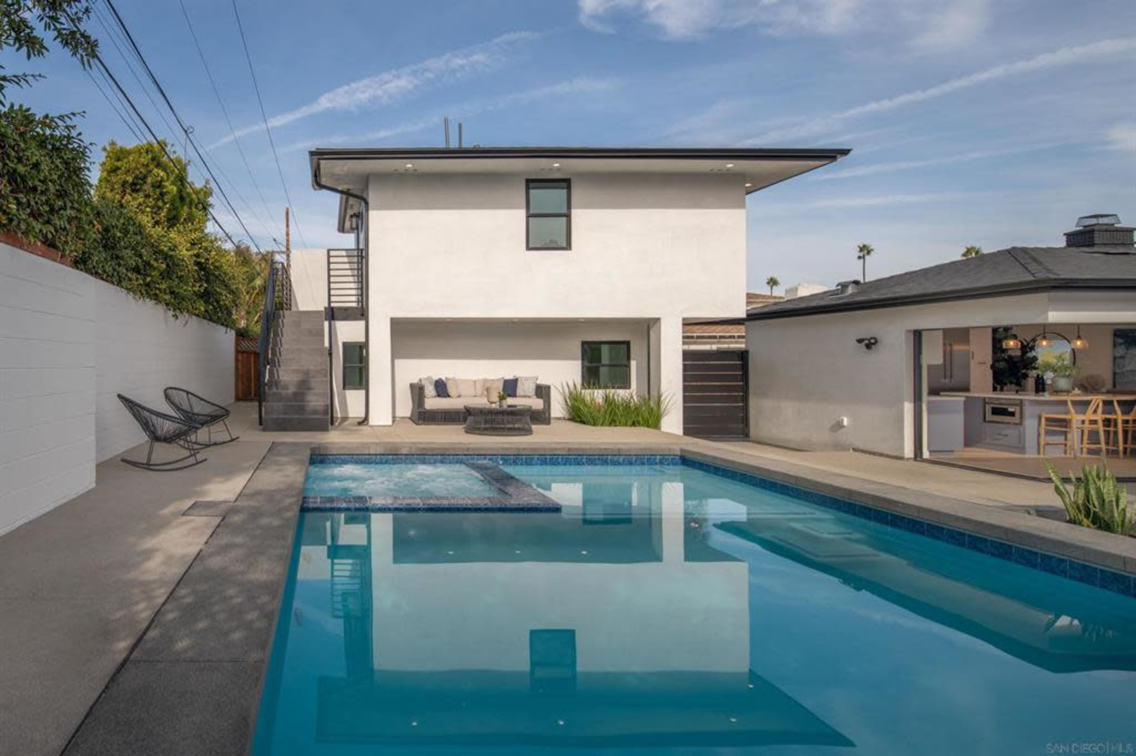 A modern house with a balcony overlooks a swimming pool surrounded by a patio and landscaped yard.