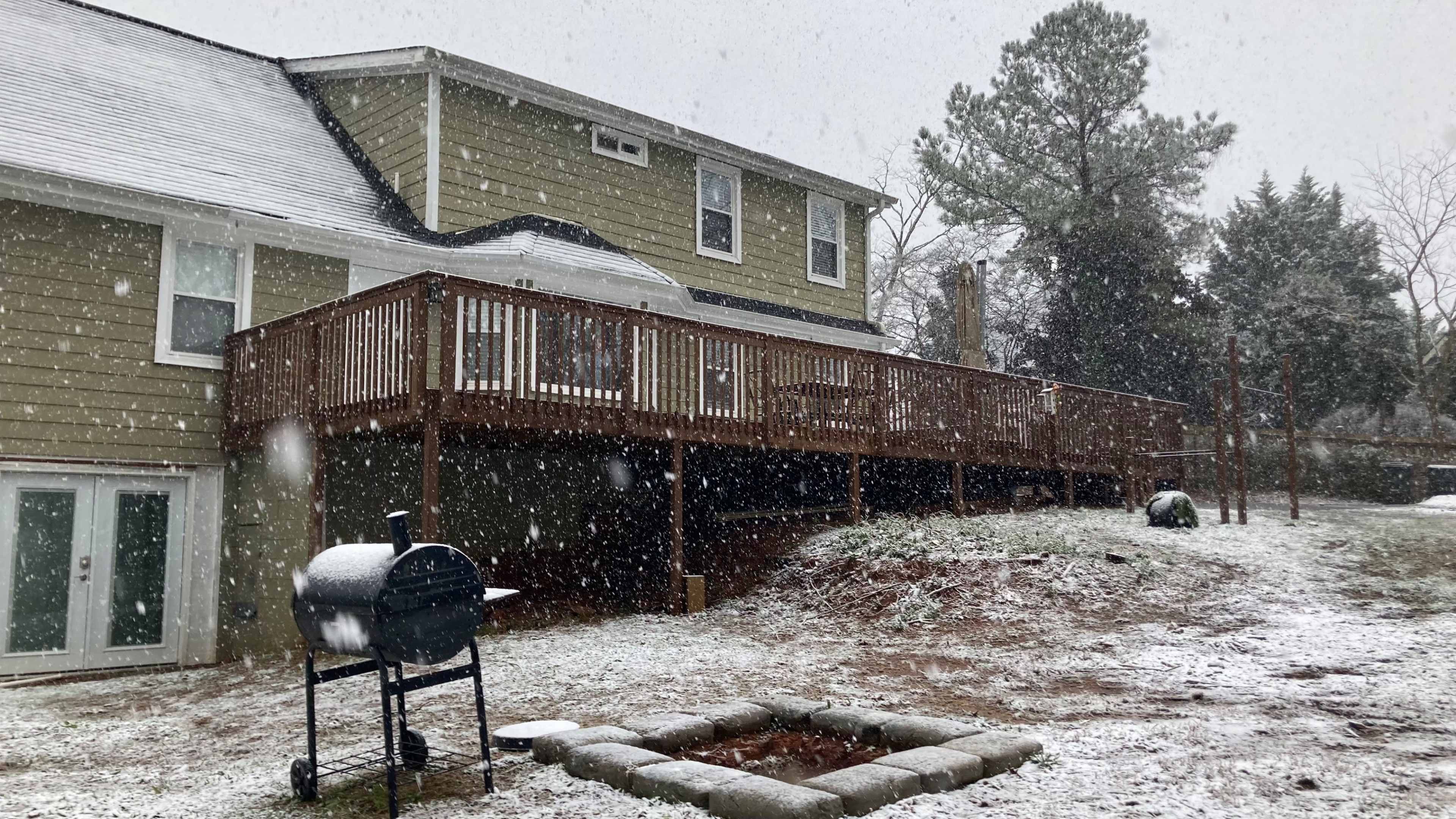 A wooden deck extends from a two-story house in a snowy backyard, featuring a grill and a fire pit surrounded by snow.