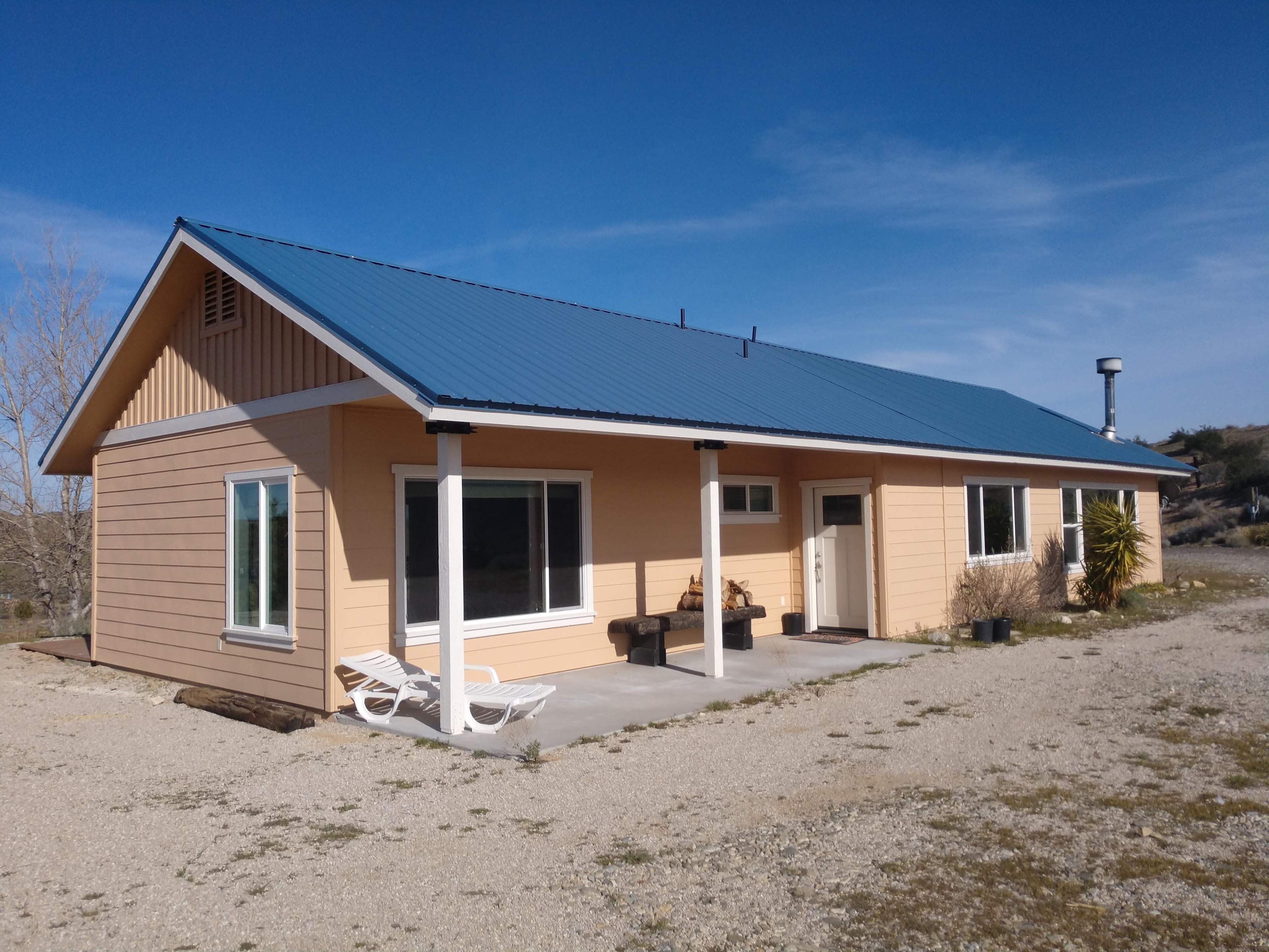 A single-story house with a blue metal roof and beige exterior sits on a gravel lot under a clear sky.