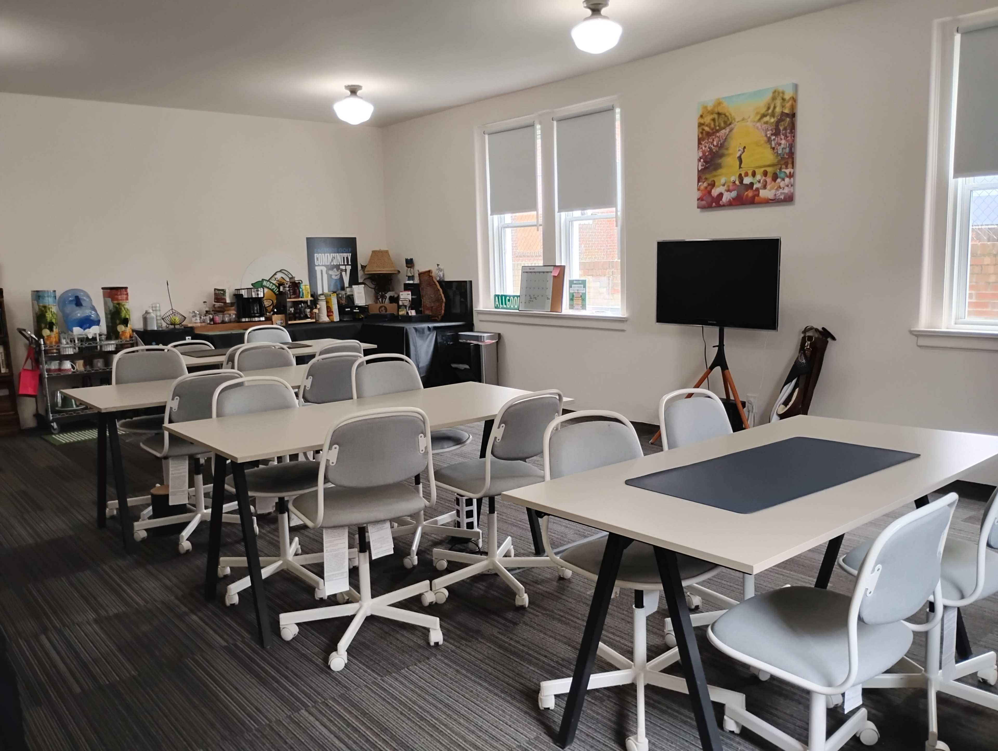 The image shows a well-lit classroom or meeting space with multiple tables and chairs arranged in a tidy manner, along with a kitchenette area and a television on the wall.