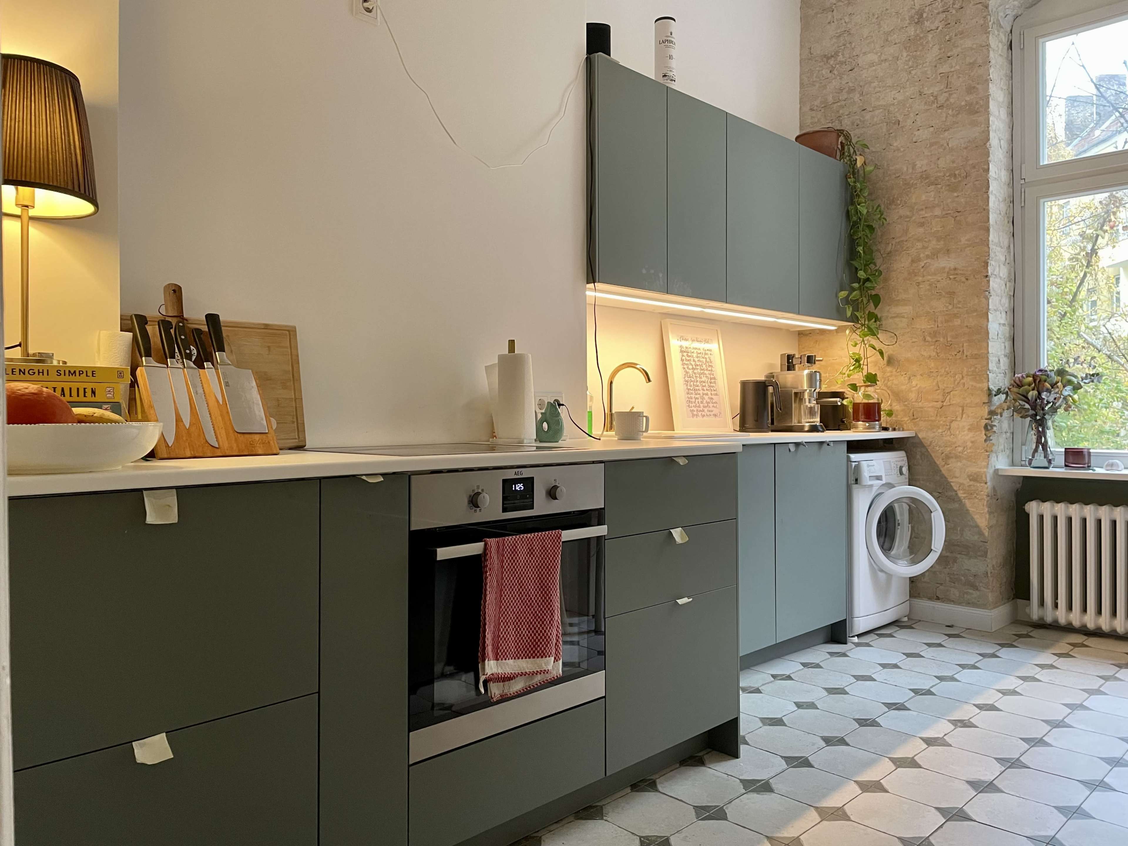 A modern kitchen with green cabinetry, a built-in oven, and a washing machine beside a window.