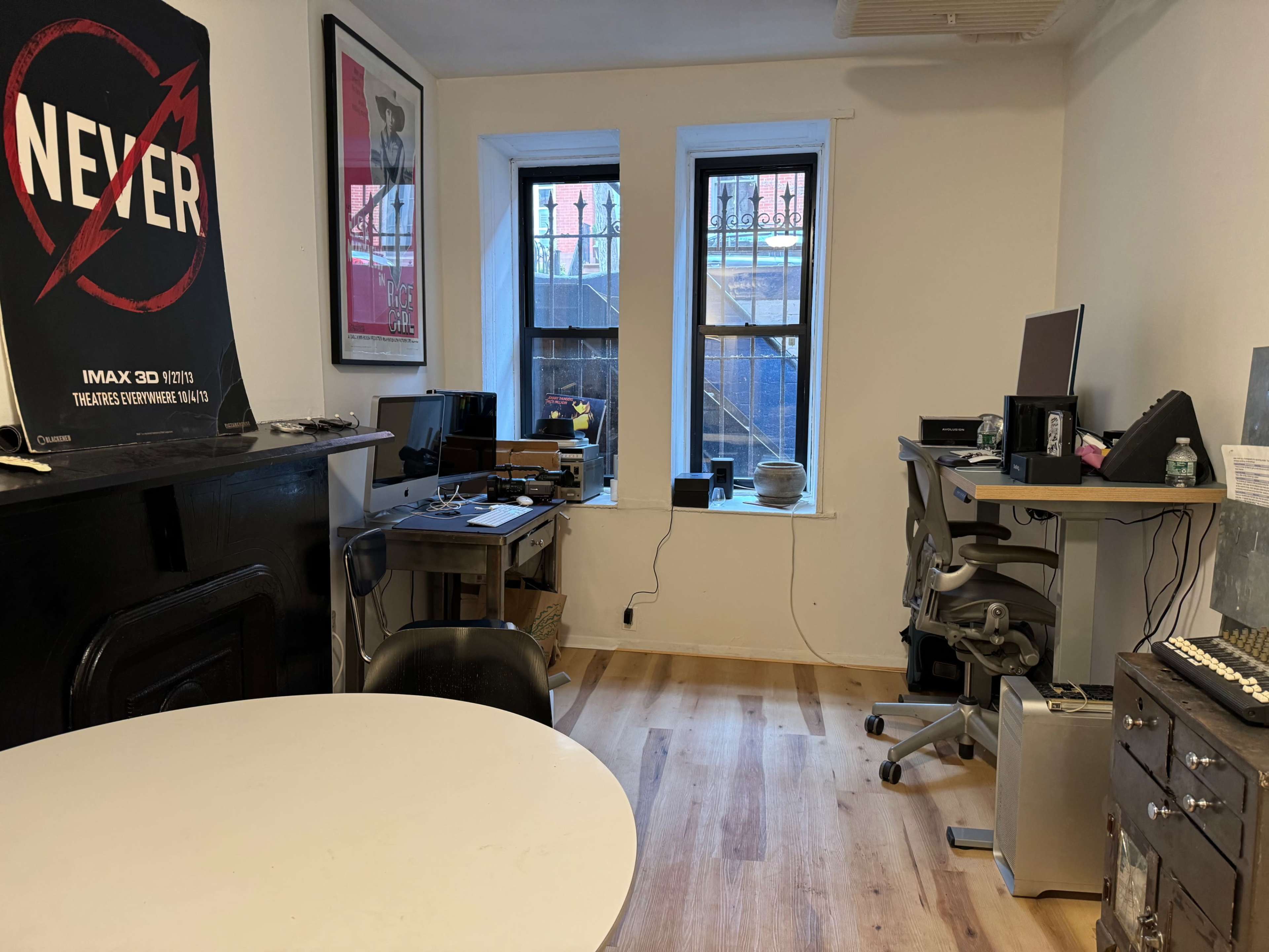 The image shows a room with two windows, a desk with a computer setup, a round table, and a black-painted fireplace, all featuring wooden flooring.