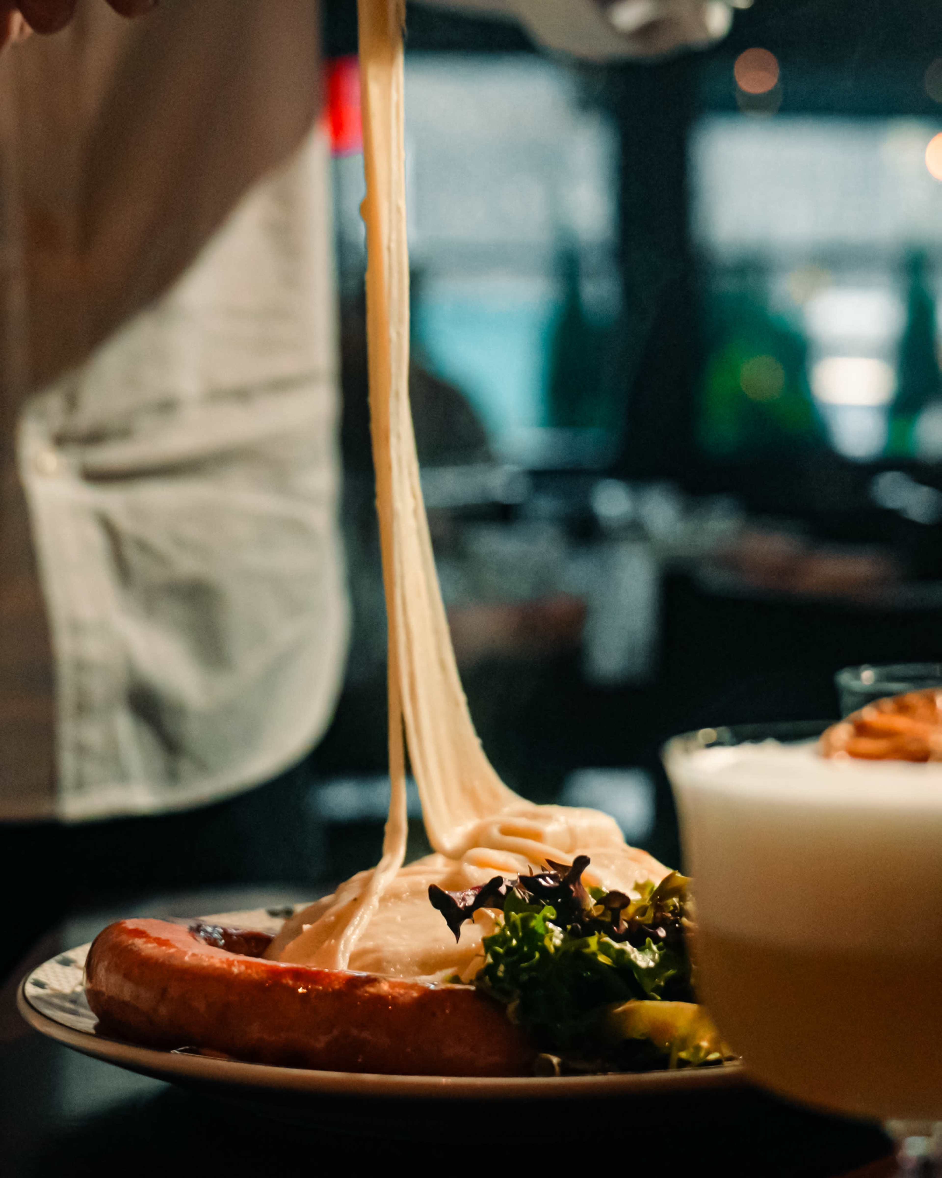A waiter drizzles cheese over a dish garnished with greens and pickles in a restaurant setting.