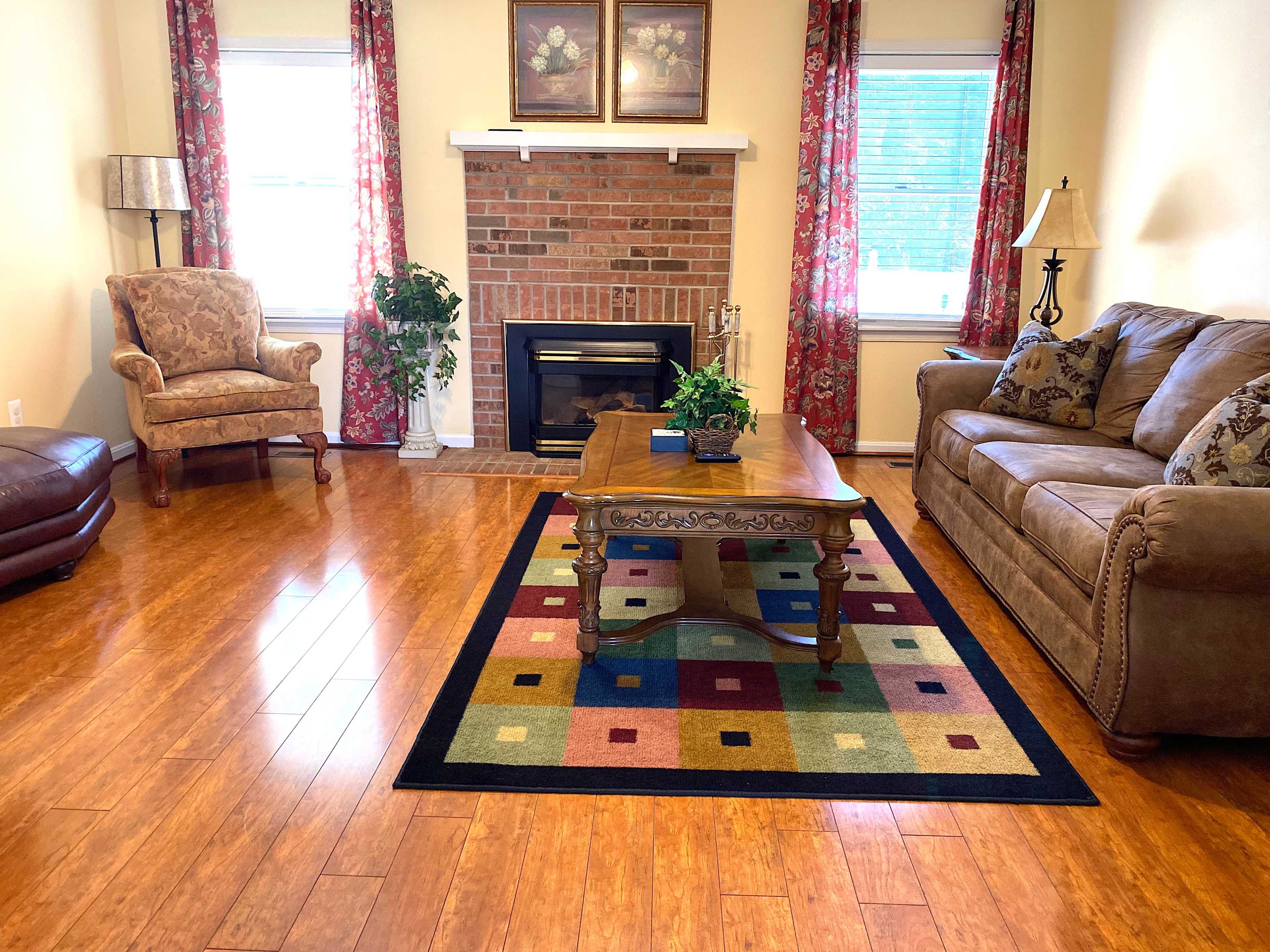 The living room features a brown leather couch, an armchair, a wooden coffee table, and a brick fireplace, all arranged on a colorful rug.