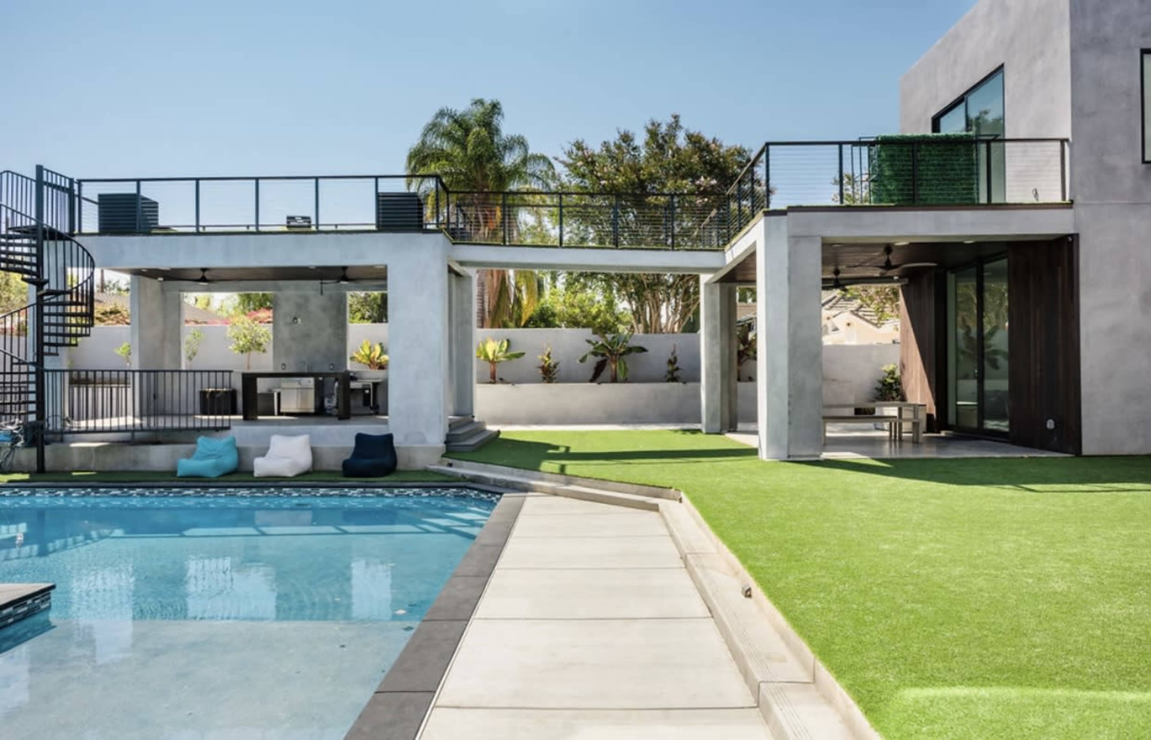 A modern backyard featuring a pool surrounded by artificial grass, with a patio area and a multi-level structure in the background.