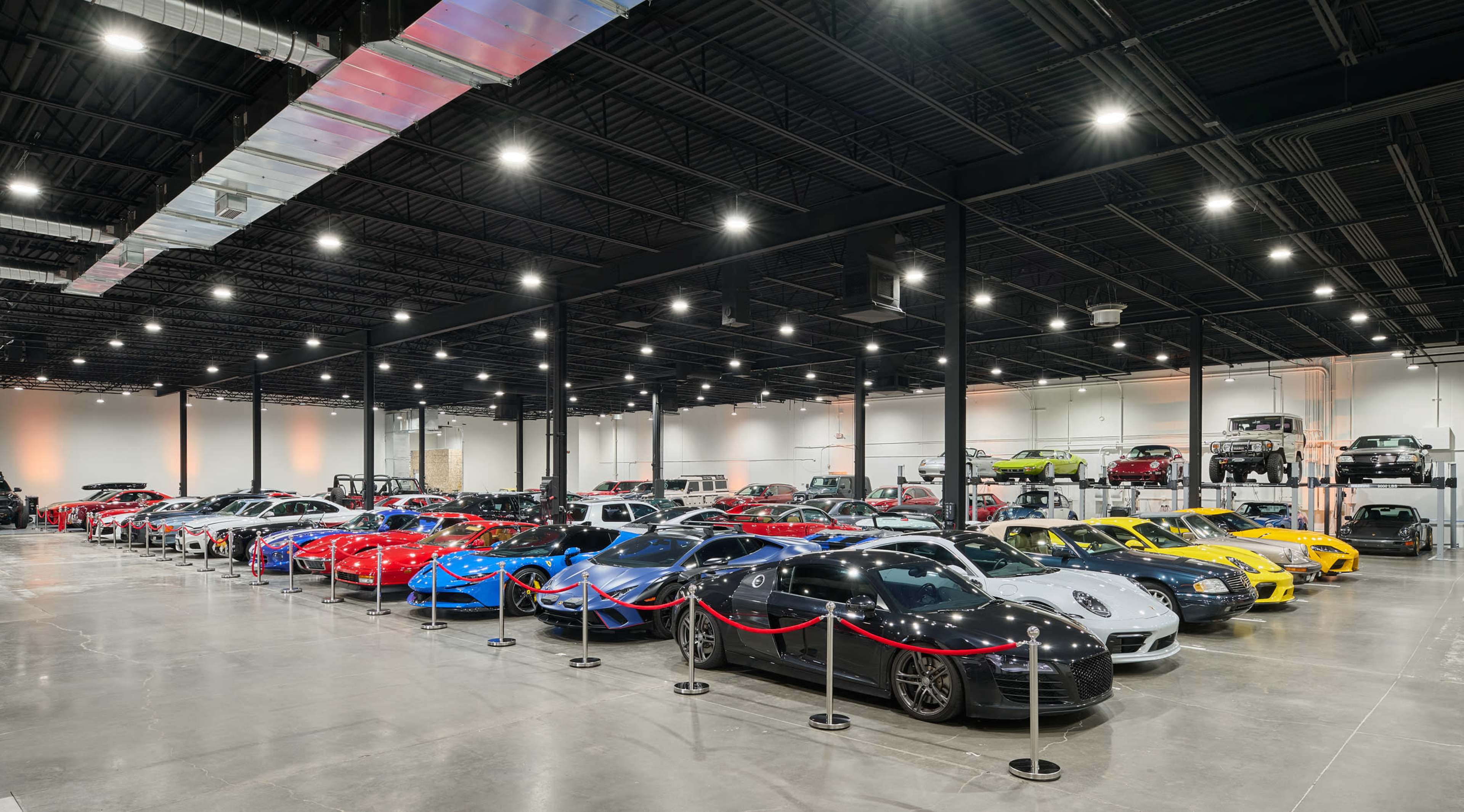The image shows a spacious car showroom featuring a variety of luxury cars lined up on the floor, separated by rope barriers.