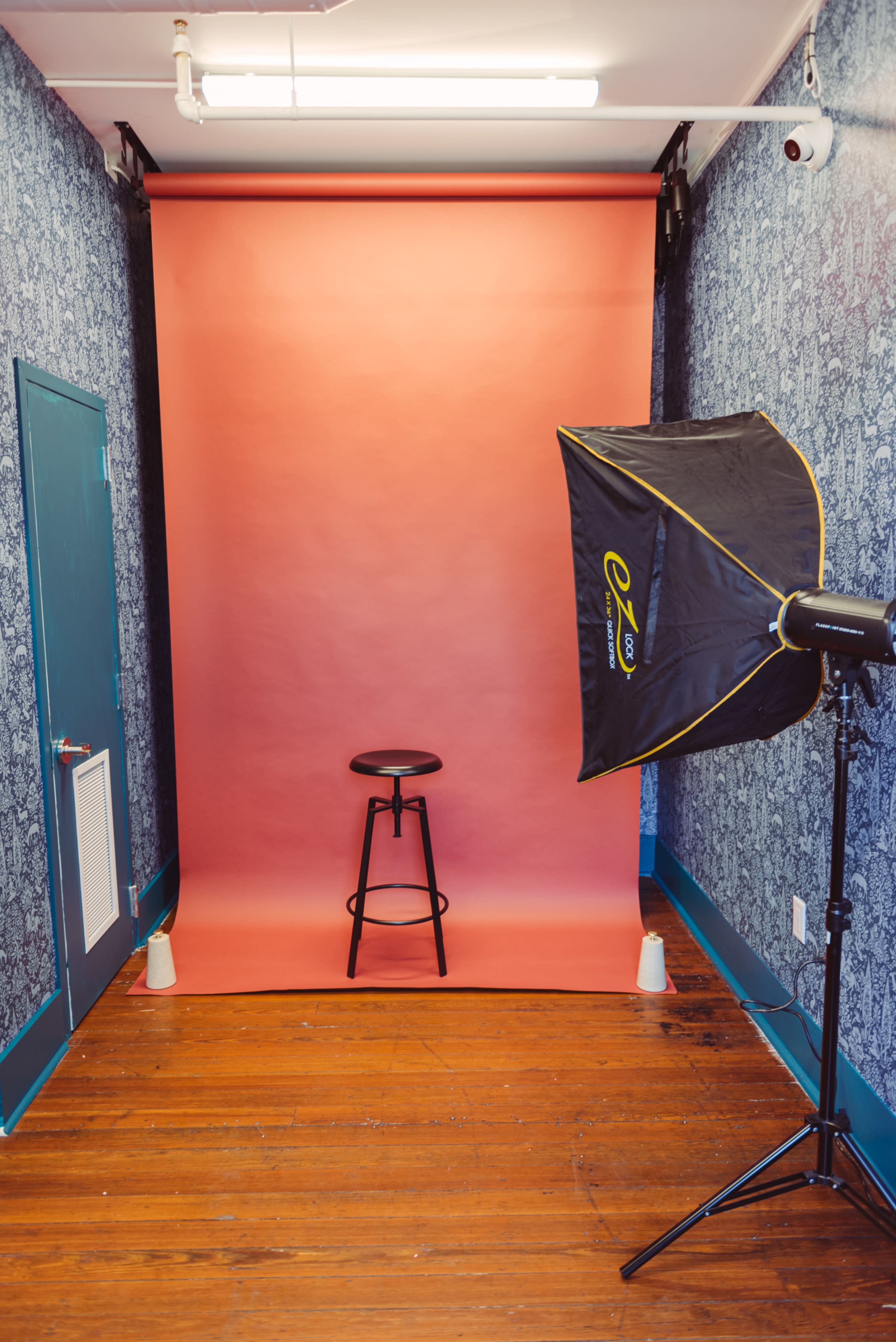A photography setup featuring a pink backdrop, a black stool in front of it, and a lighting umbrella on the side.