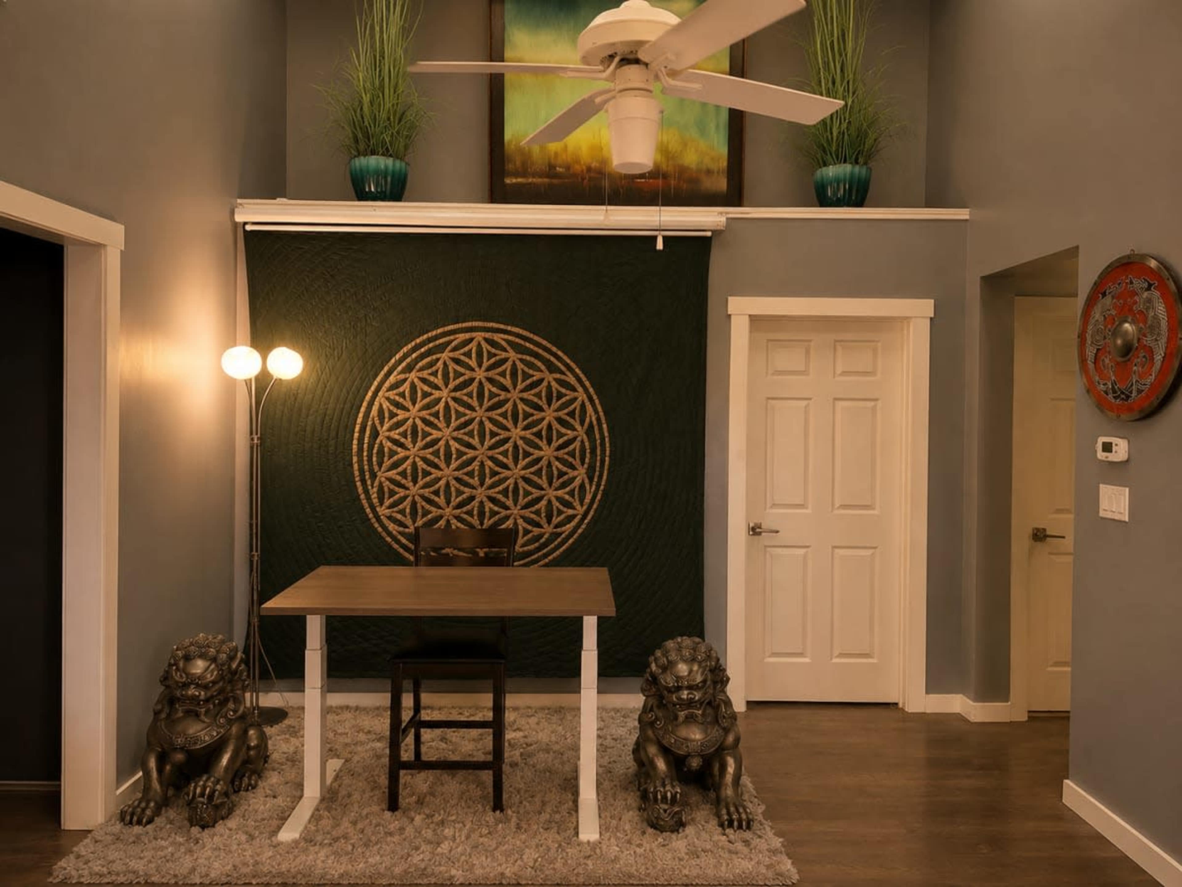 The image shows a modern room with a small table and chair in front of a decorative green wall hanging, flanked by two lion sculptures and featuring a ceiling fan above.
