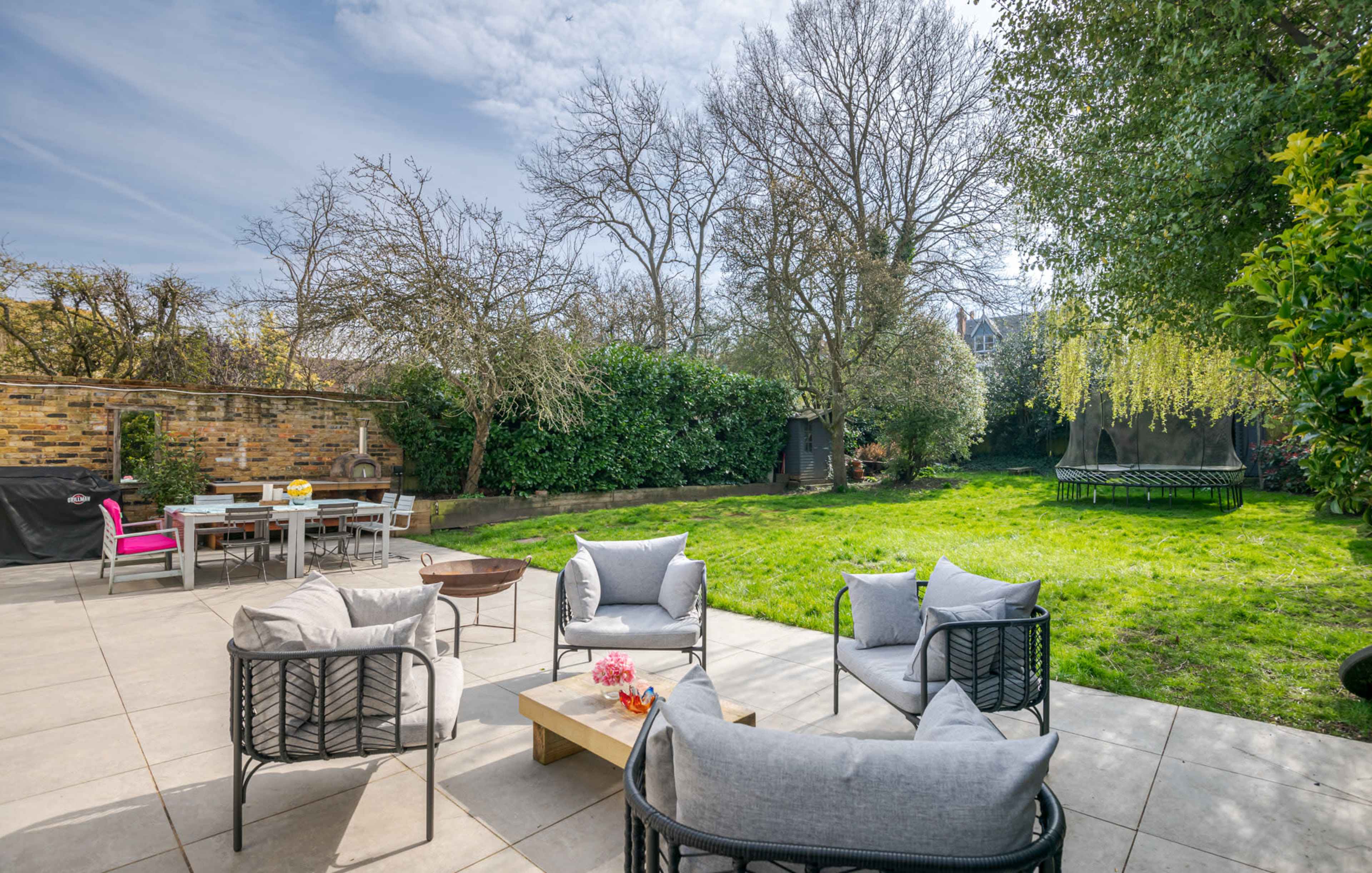 A spacious backyard features a seating area with plush chairs, a wooden coffee table, and a dining table surrounded by trees and greenery.