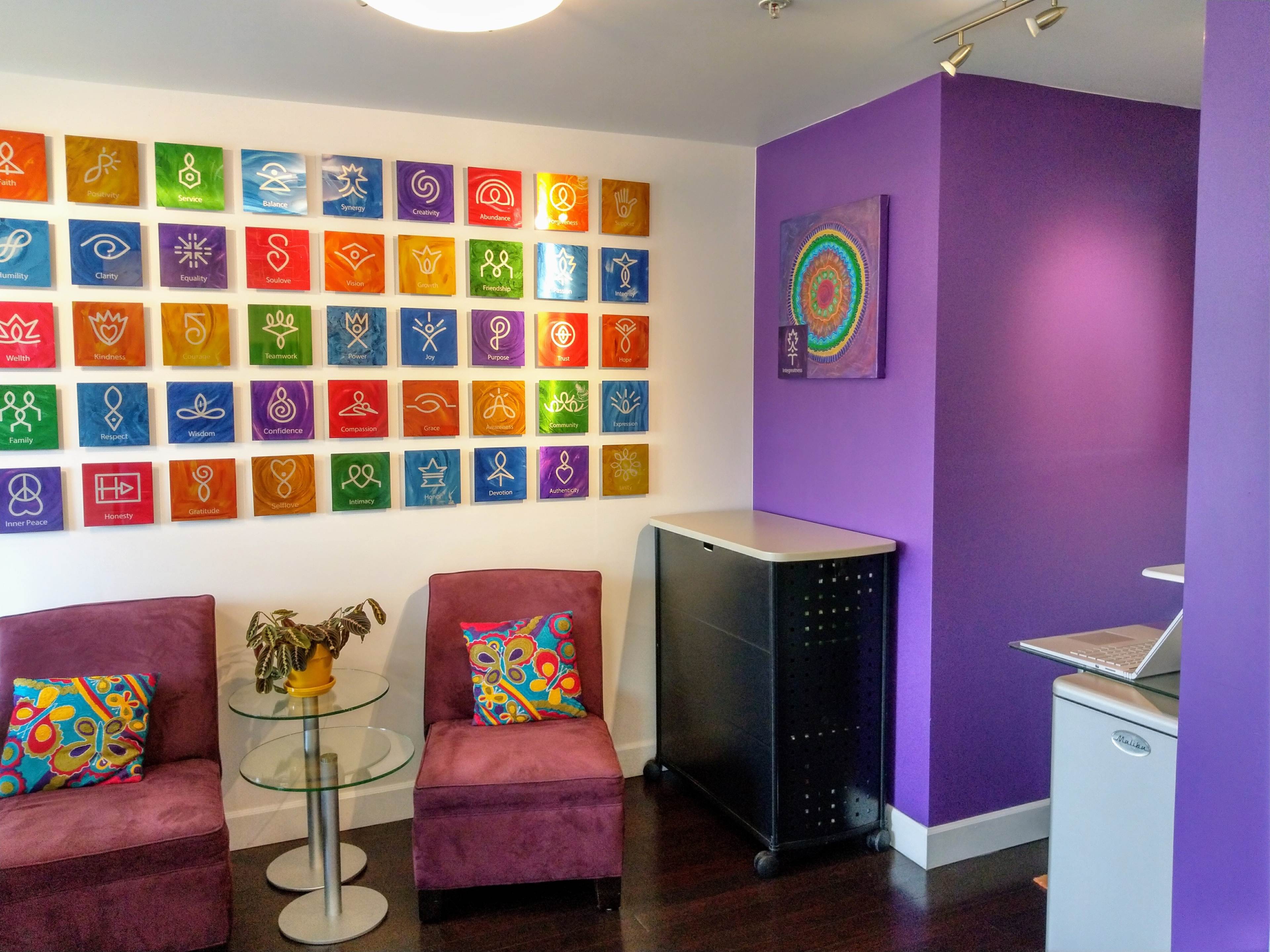 A brightly colored waiting area with purple walls, two plush chairs, a glass table, and a display of artwork mounted on the wall.