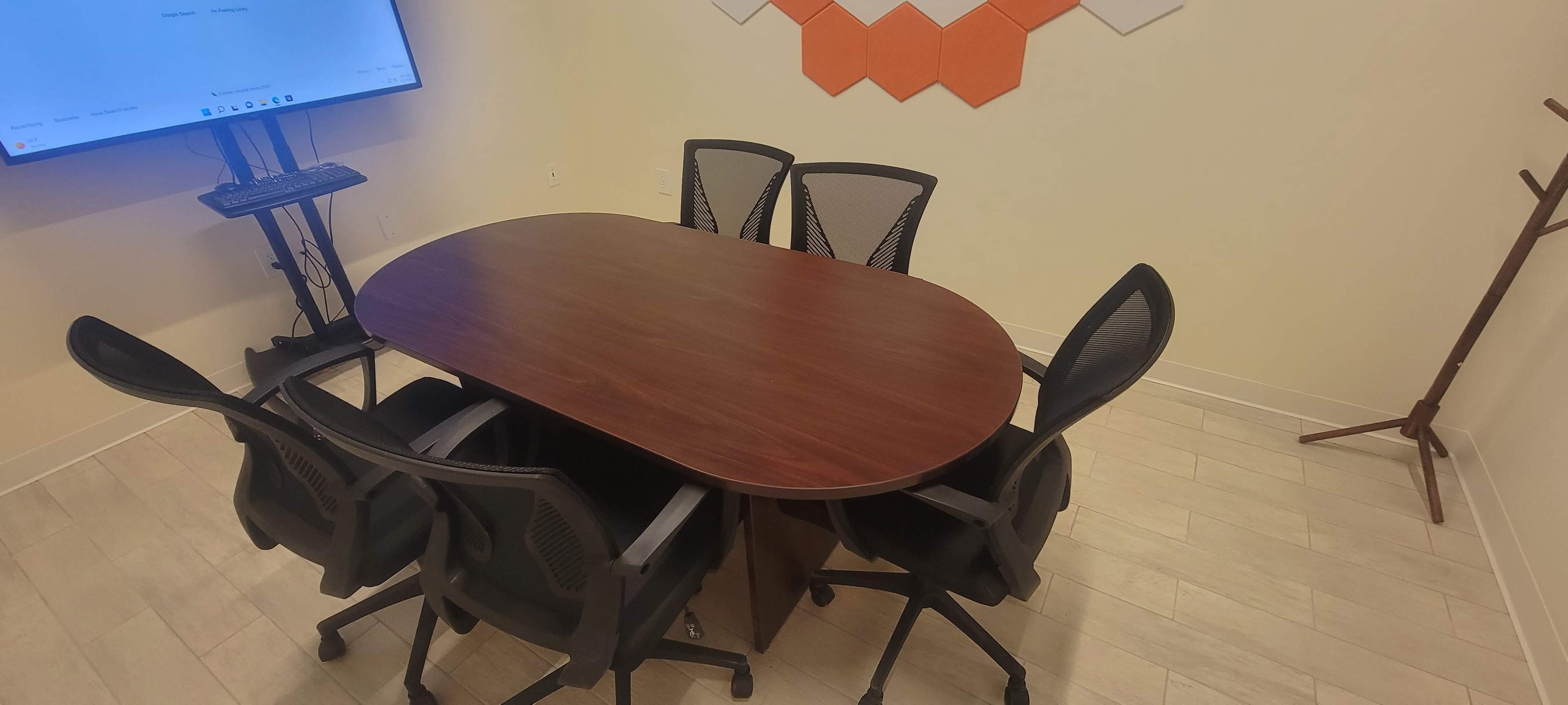 A conference room features a rounded wooden table surrounded by five black office chairs, with a wall-mounted screen and a coat rack nearby.