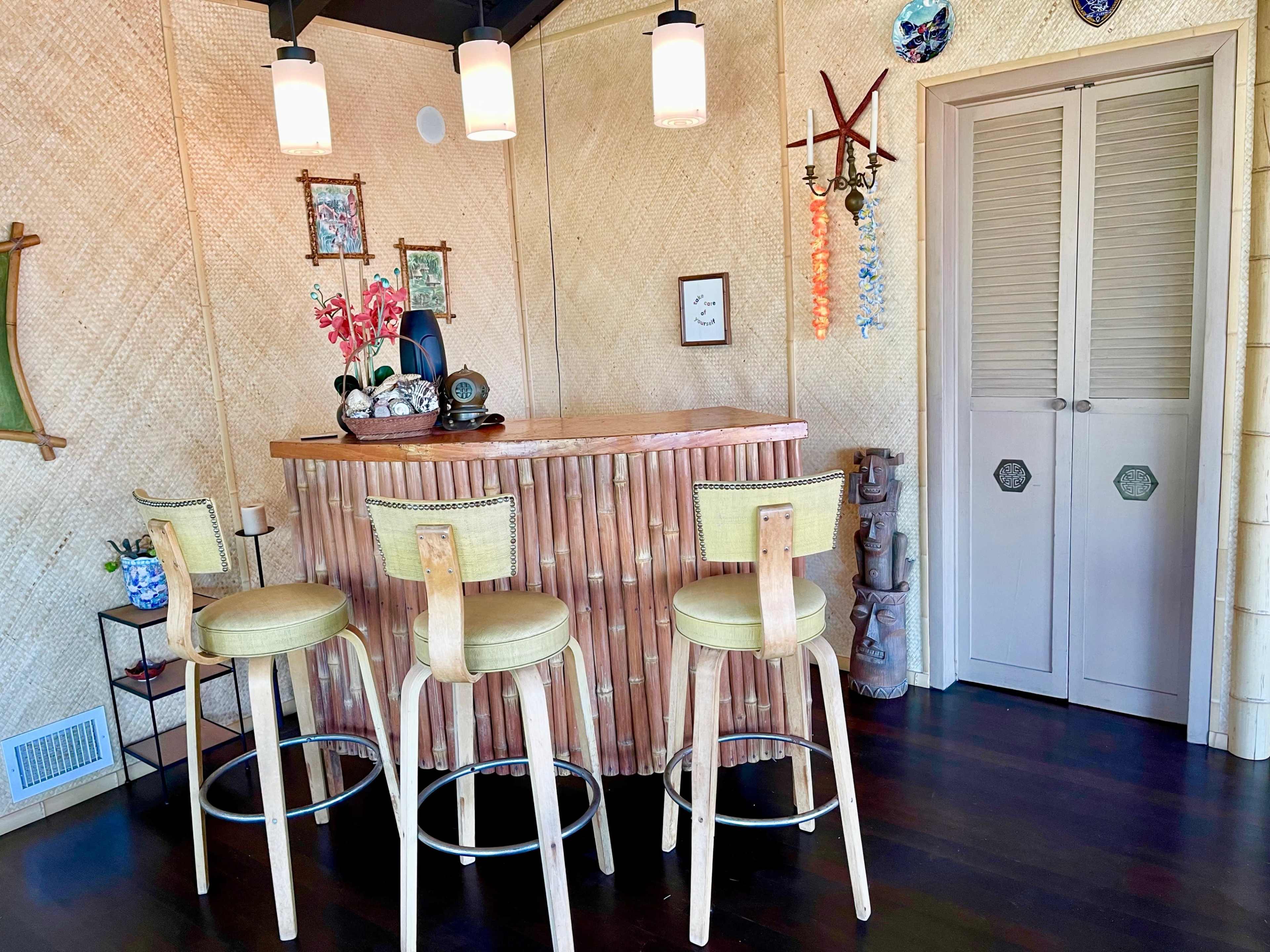 A bamboo-themed bar area with three wooden stools, decorative wall art, and a closed closet in a room with woven walls.