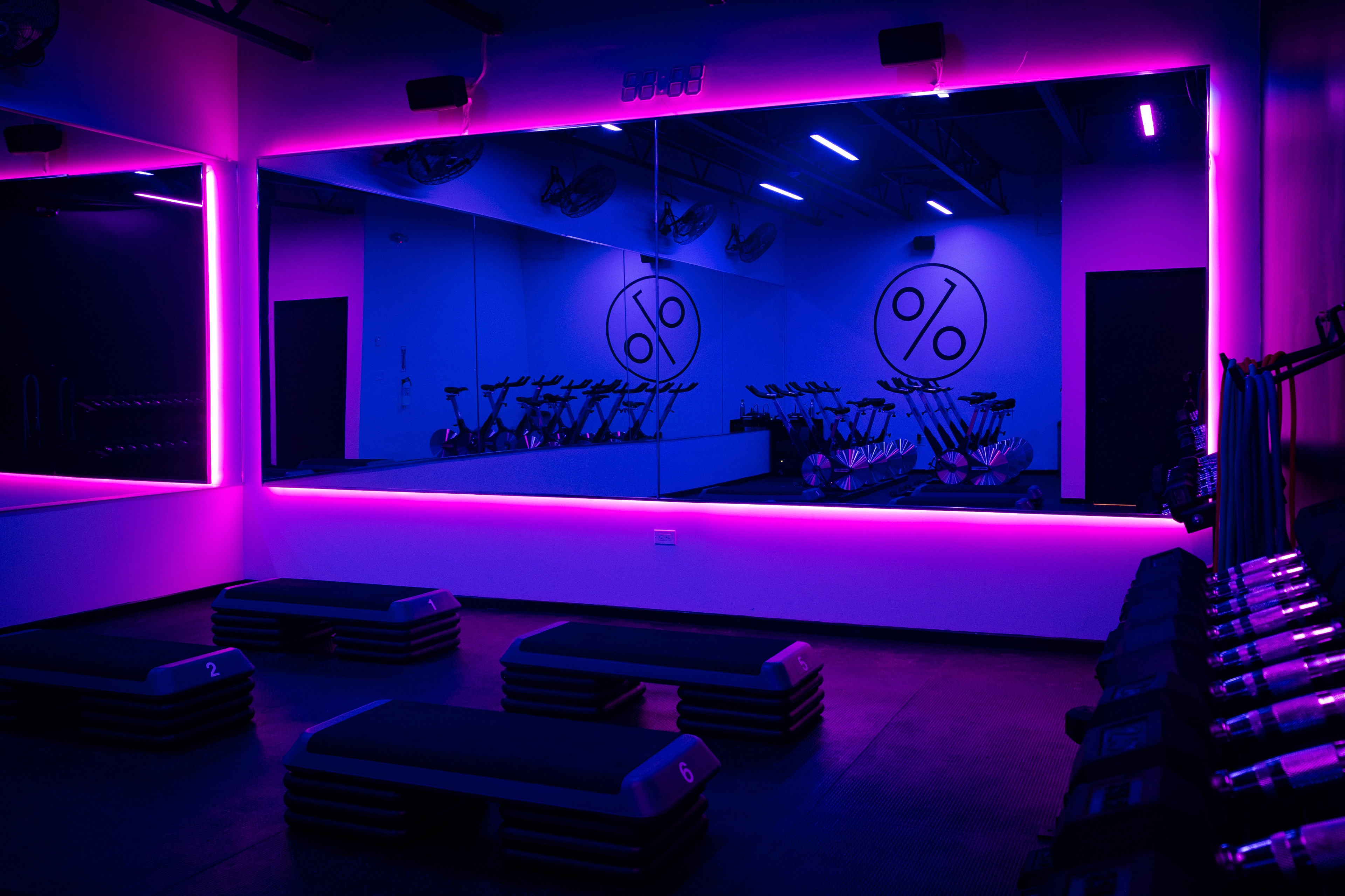 A brightly lit gym area features exercise benches and spinning bikes, with vivid purple lighting illuminating the space and a mirrored wall displaying a percentage symbol.