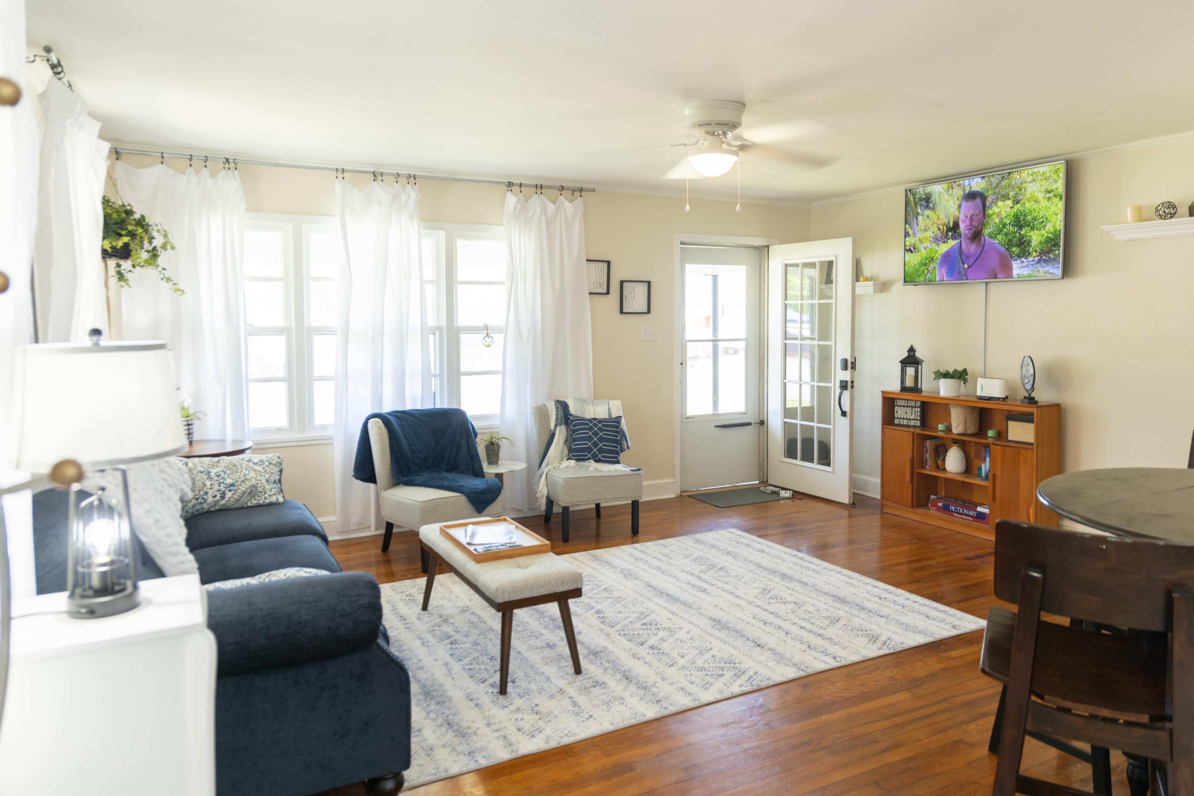 A living room features a sofa, two armchairs, a coffee table, and a television mounted on the wall, with large windows allowing natural light and white curtains draping beside them.