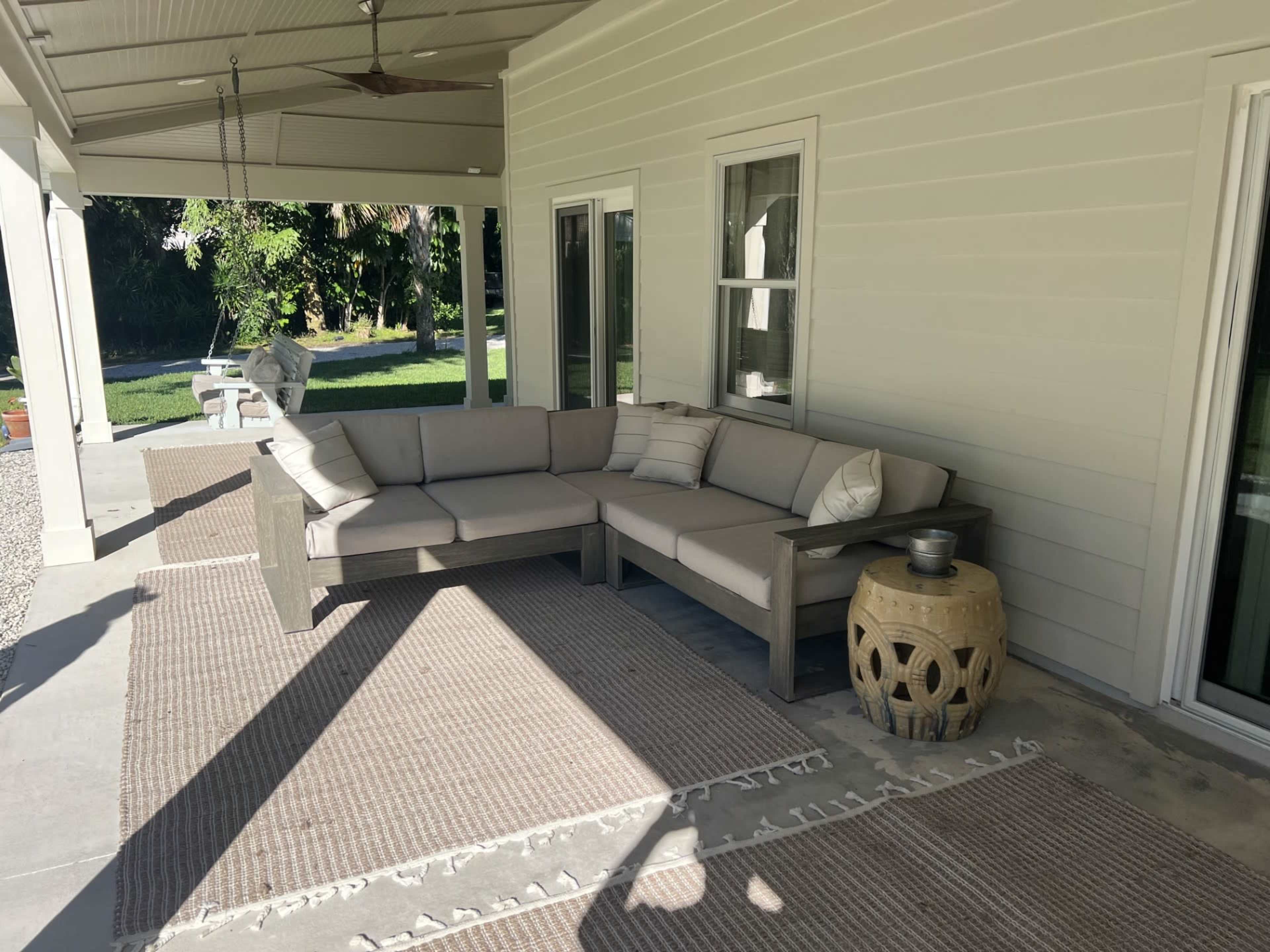 A sectional outdoor sofa with light-colored cushions is positioned on a covered patio, accompanied by a round wooden table and decorative rugs.