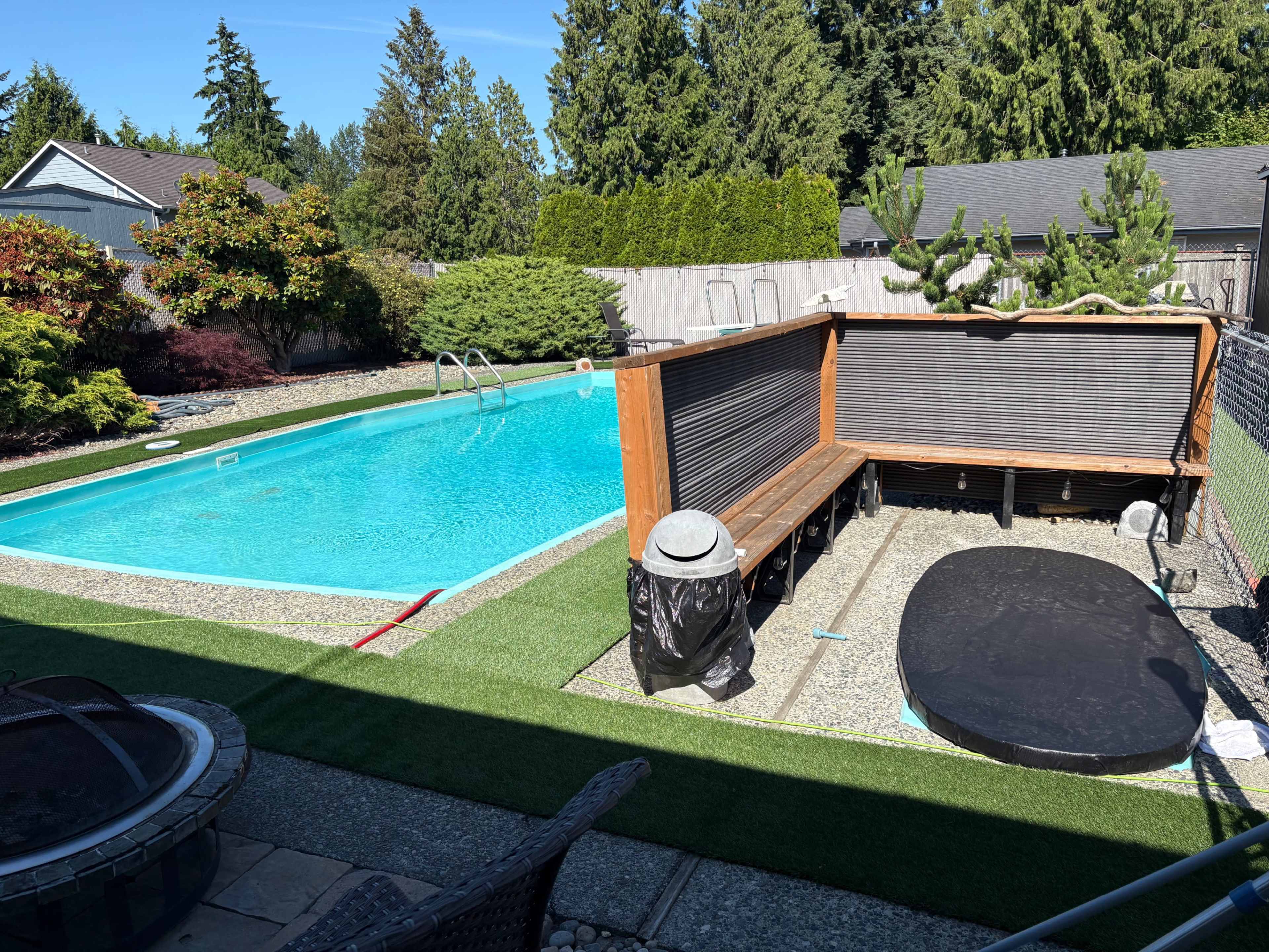 The image shows a backyard scene featuring a clear blue swimming pool surrounded by landscaped greenery and a wooden seating area.
