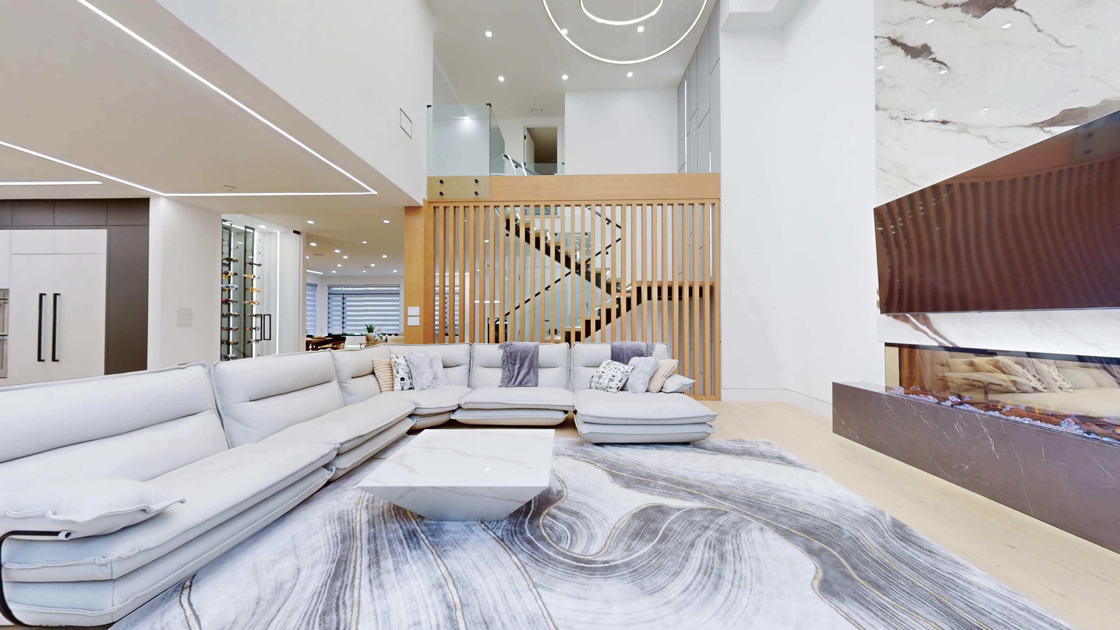 A modern living room features a large sectional sofa, a marble coffee table, and a decorative rug, with a staircase visible in the background.