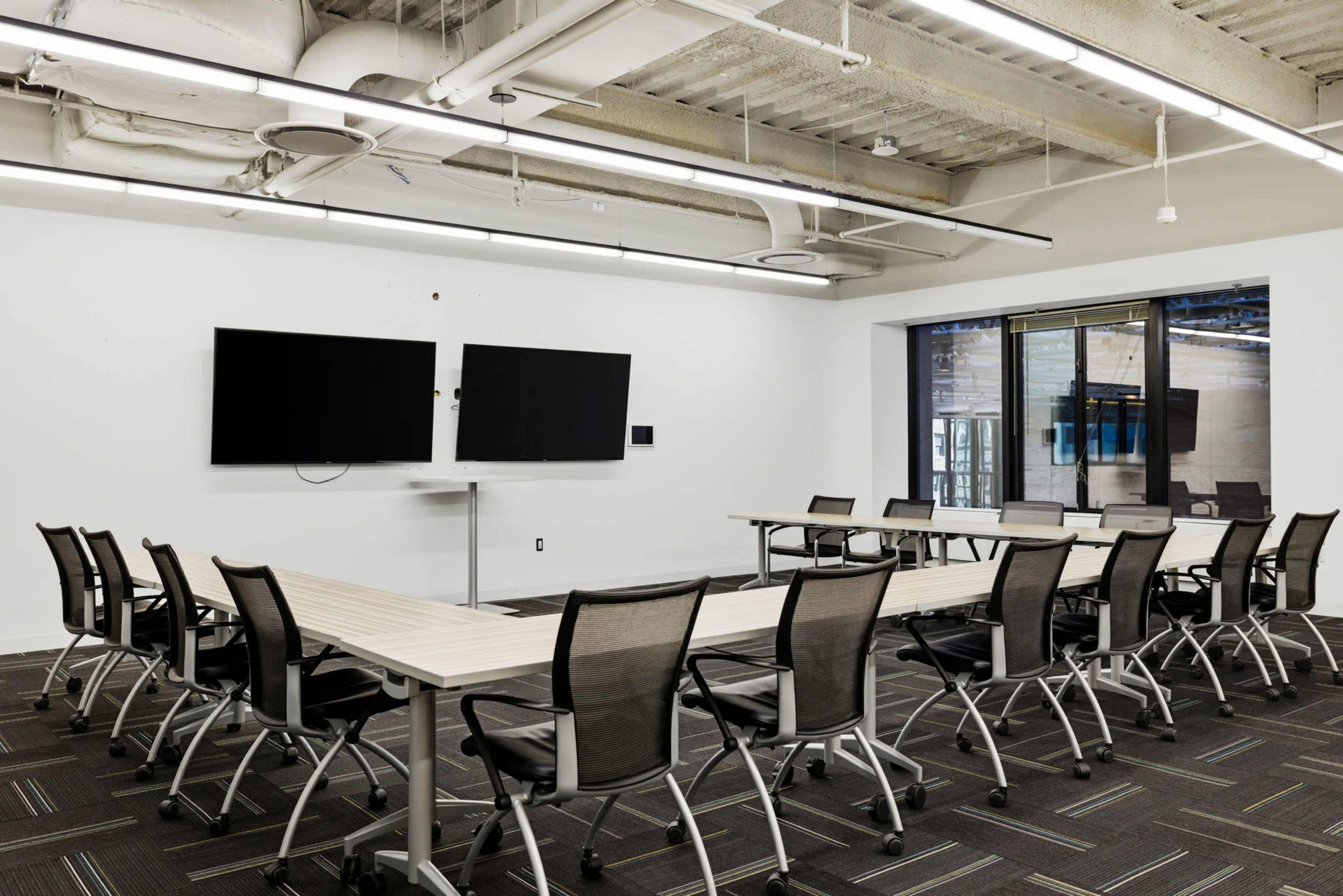 The image shows a modern conference room with two wall-mounted screens, a long table surrounded by chairs, and large windows providing natural light.