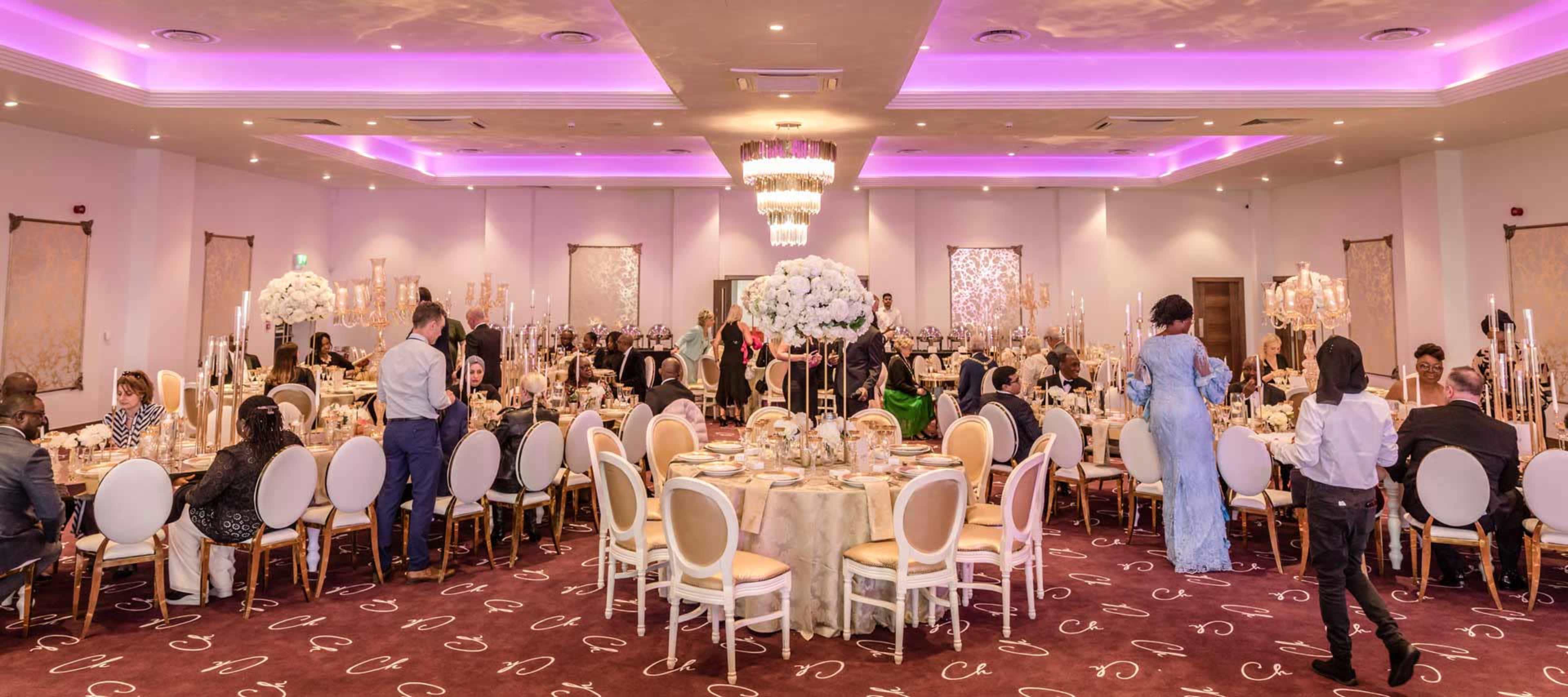 A banquet hall is set up for an elegant event, featuring round tables adorned with floral centerpieces and guests mingling throughout the space.