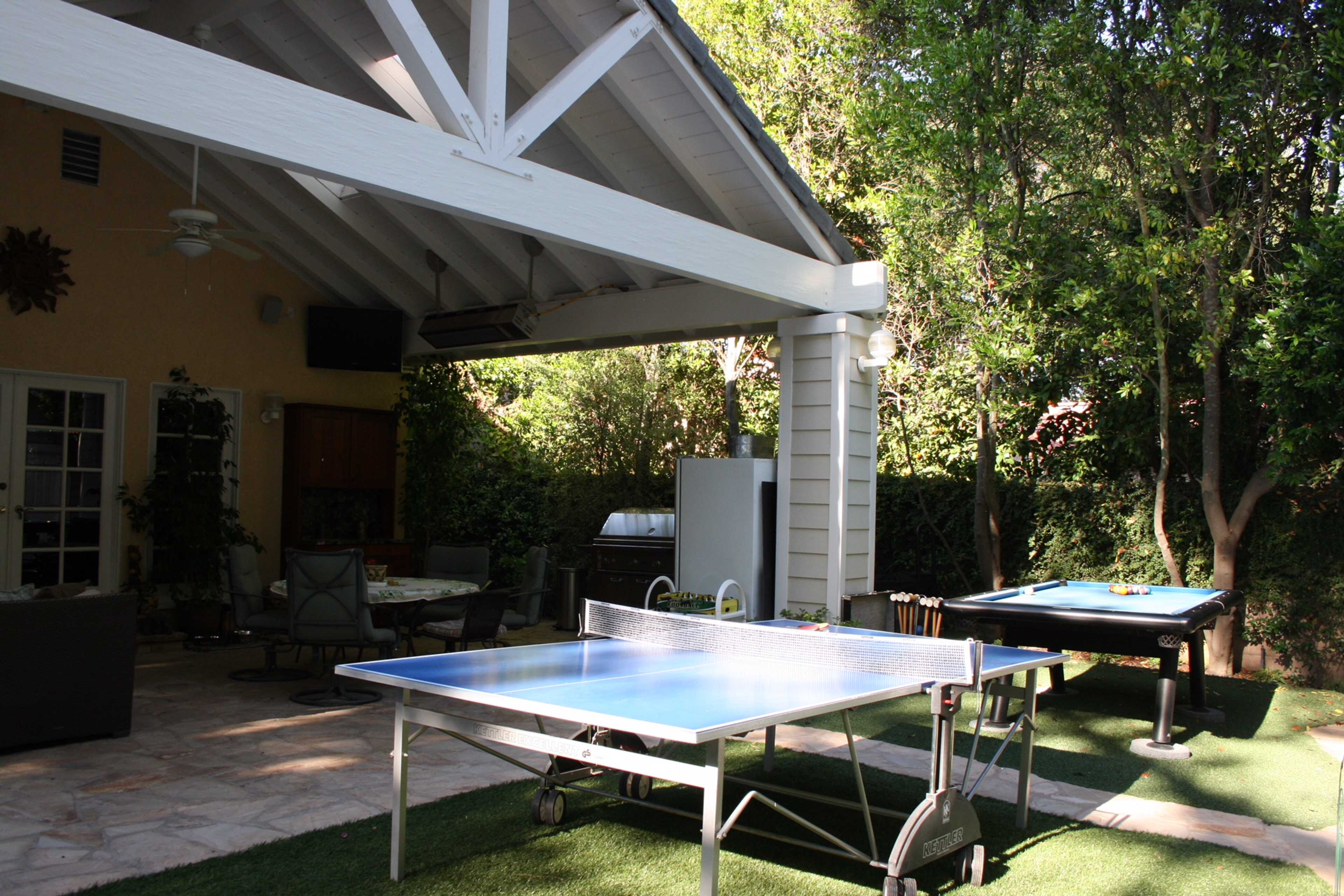 A covered patio area with two ping pong tables set on a grassy surface, surrounded by trees.