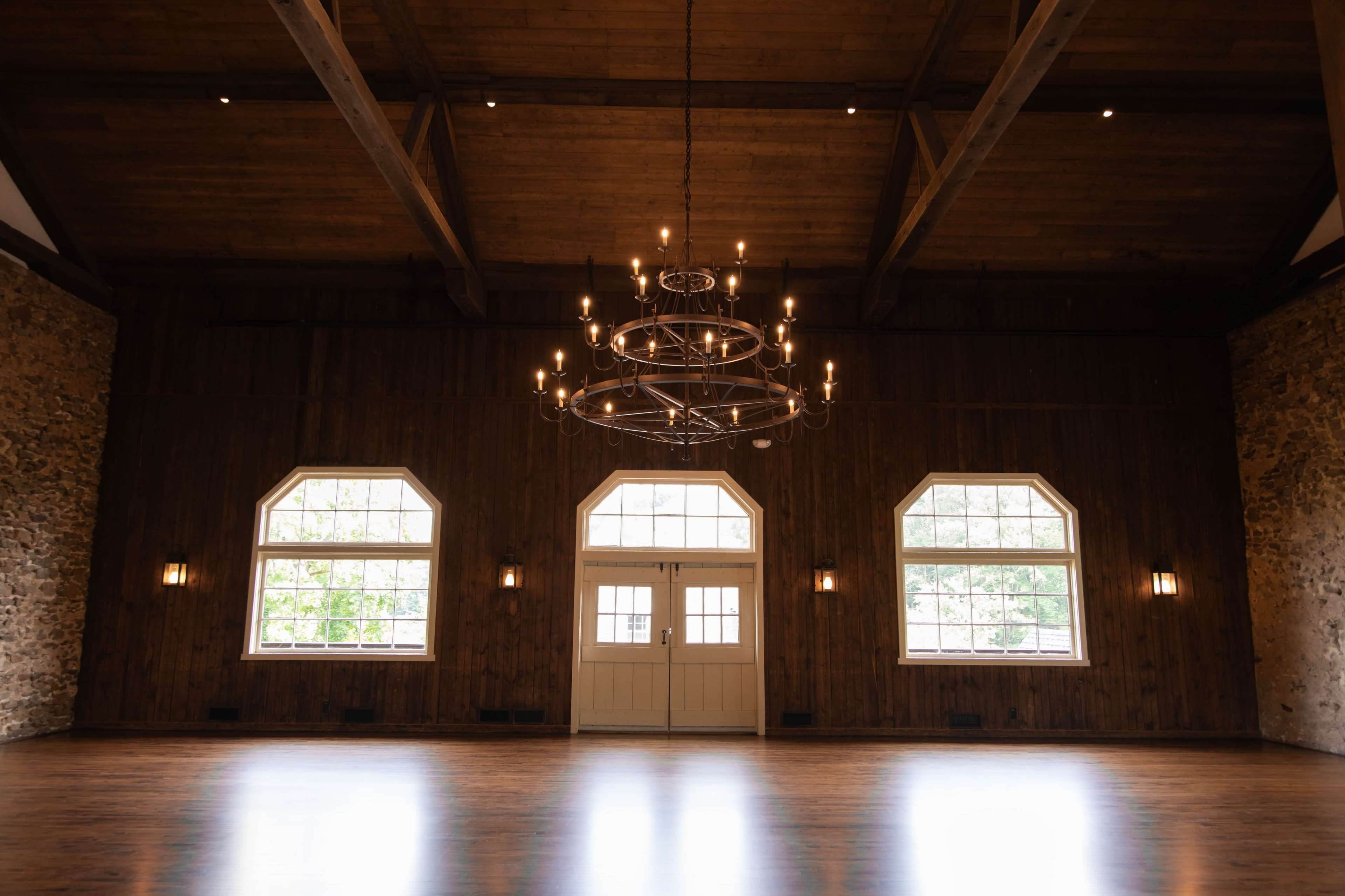 The image shows a spacious interior of a hall with wooden beams, stone walls, large windows, and a chandelier hanging from the ceiling.