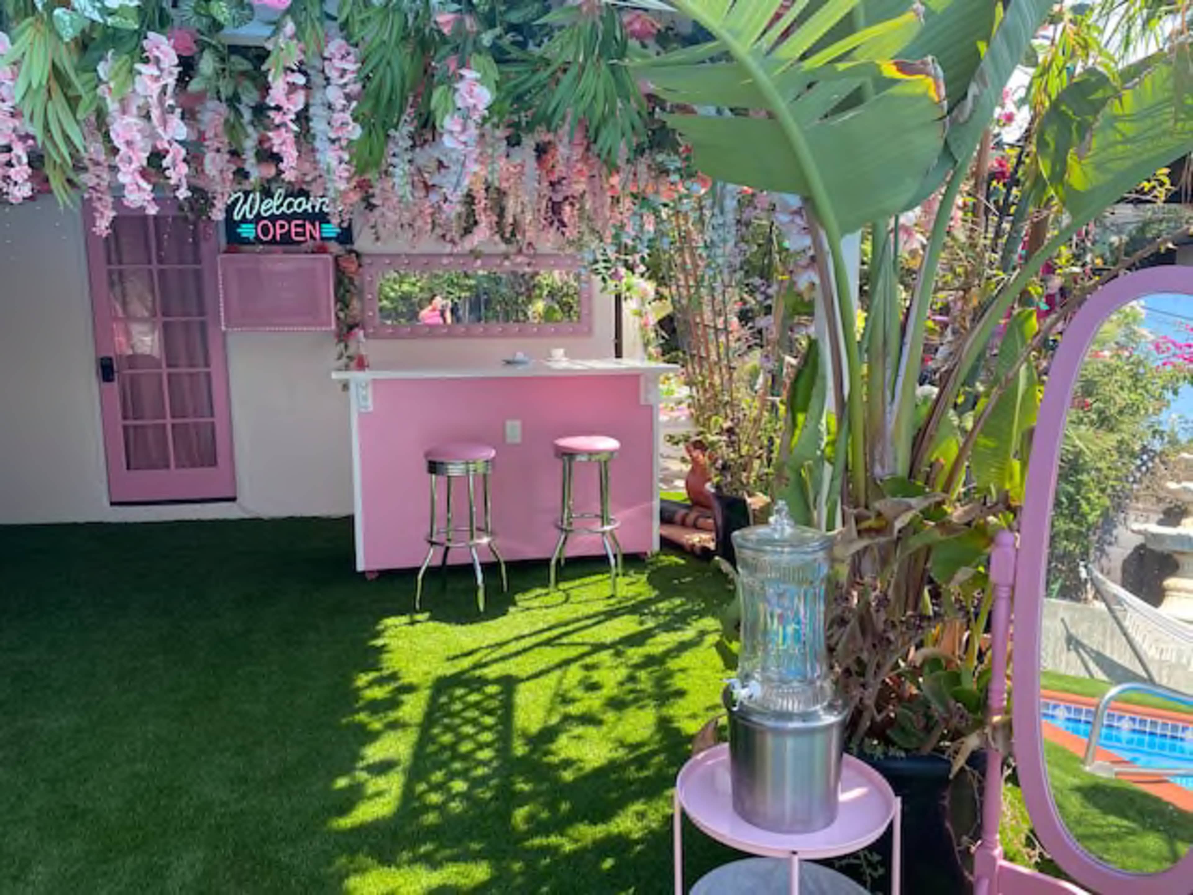 A decorated outdoor space with pink furniture, artificial flowers hanging from above, and a view of a pool in the background.