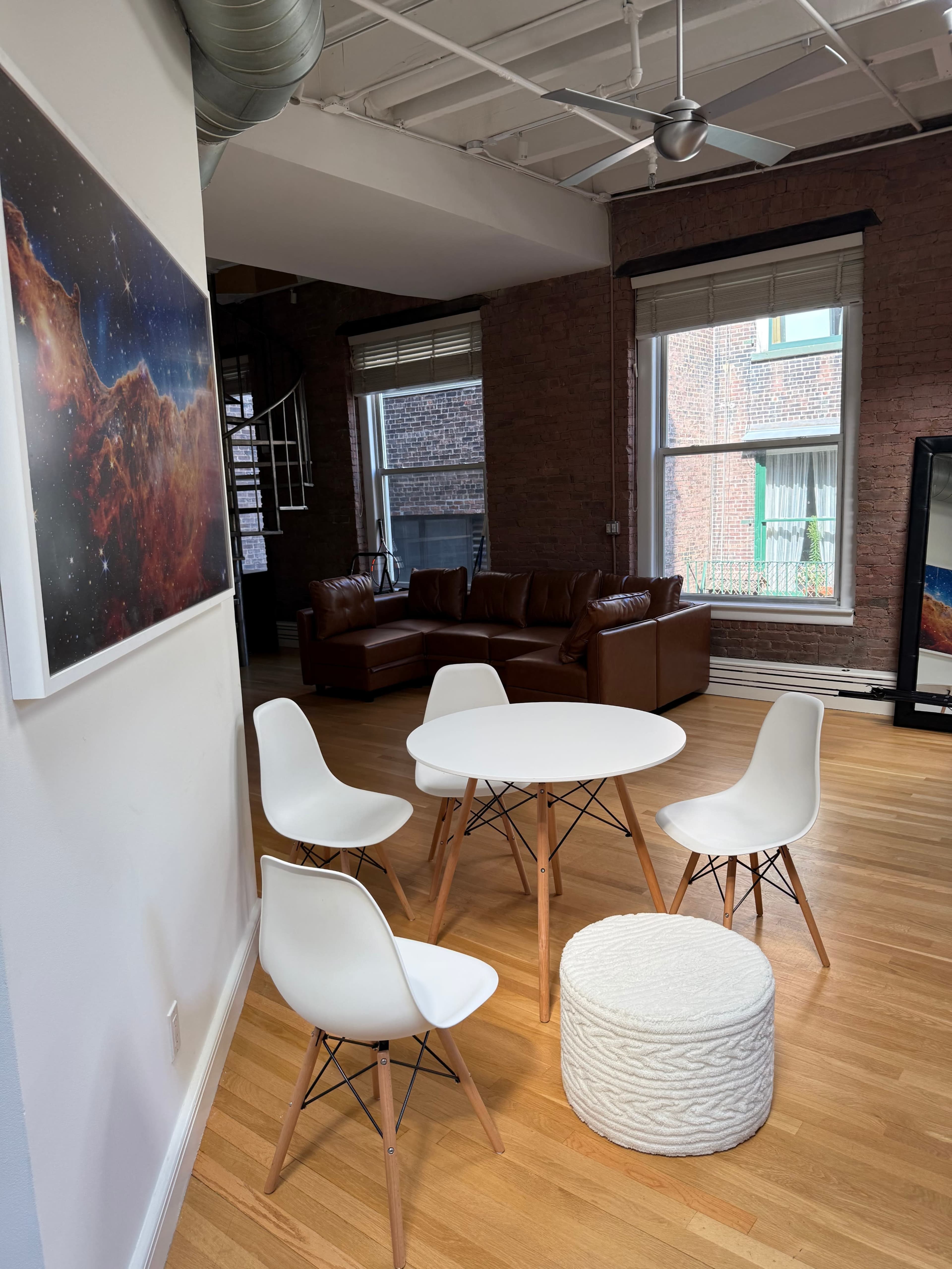 A modern interior features a round white table surrounded by four chairs and a textured ottoman, with a brown leather couch visible in the background.