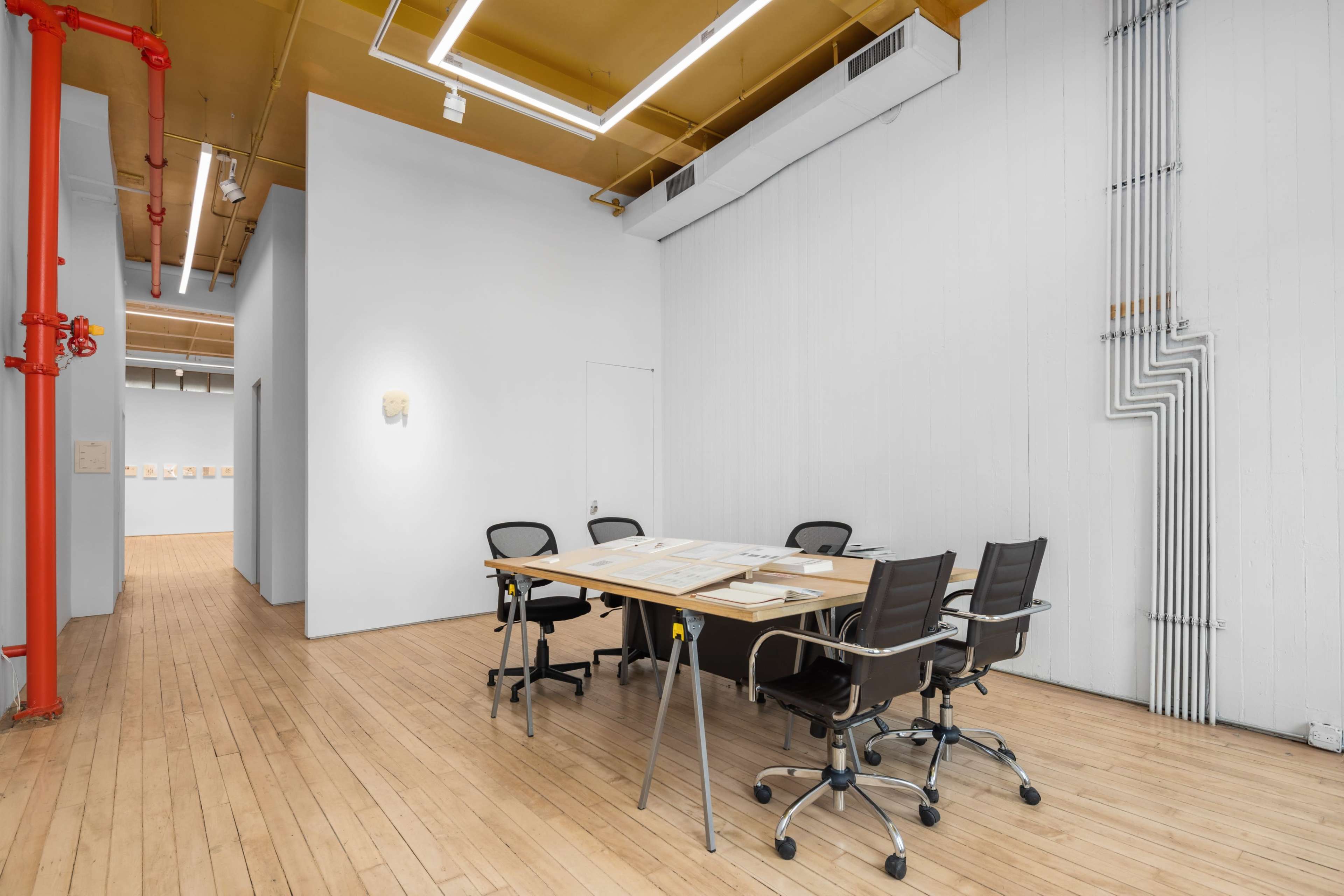 A spacious, well-lit meeting room features a wooden table surrounded by four black chairs, with white walls and visible industrial piping.
