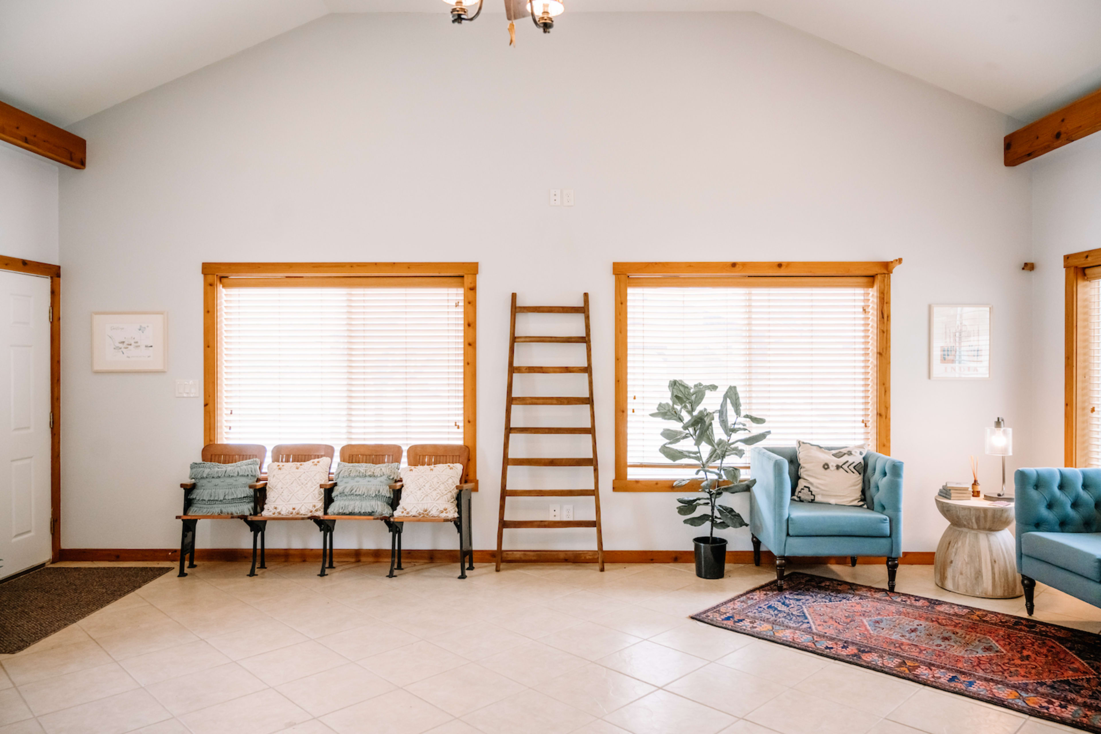 A bright, spacious room features seating along one wall, large windows with blinds, a ladder against another wall, and a potted plant beside a blue armchair and a side table.