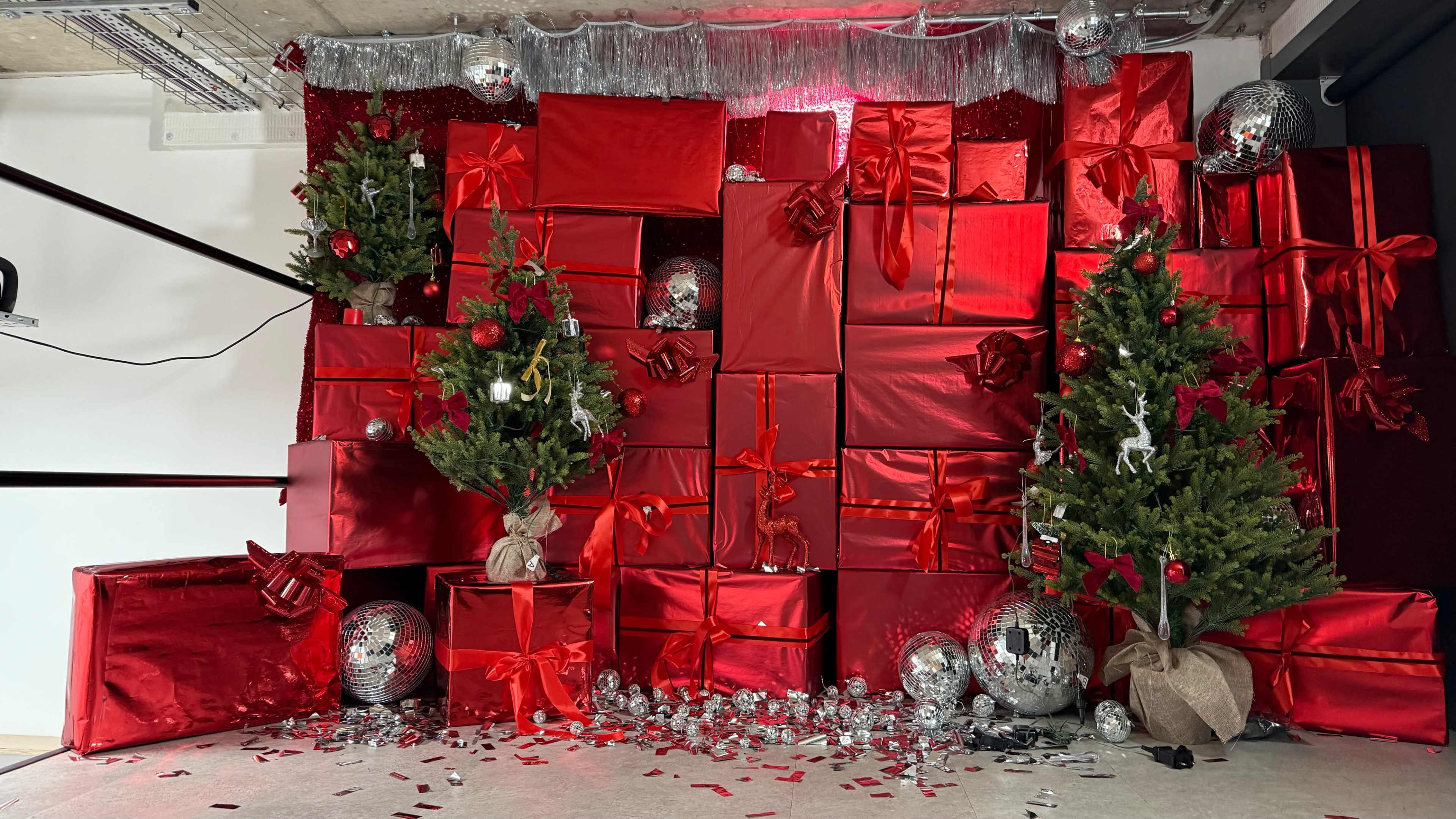 The scene features a wall covered with red gift-wrapped boxes, two small Christmas trees, and decorative ornaments, including disco balls and confetti.