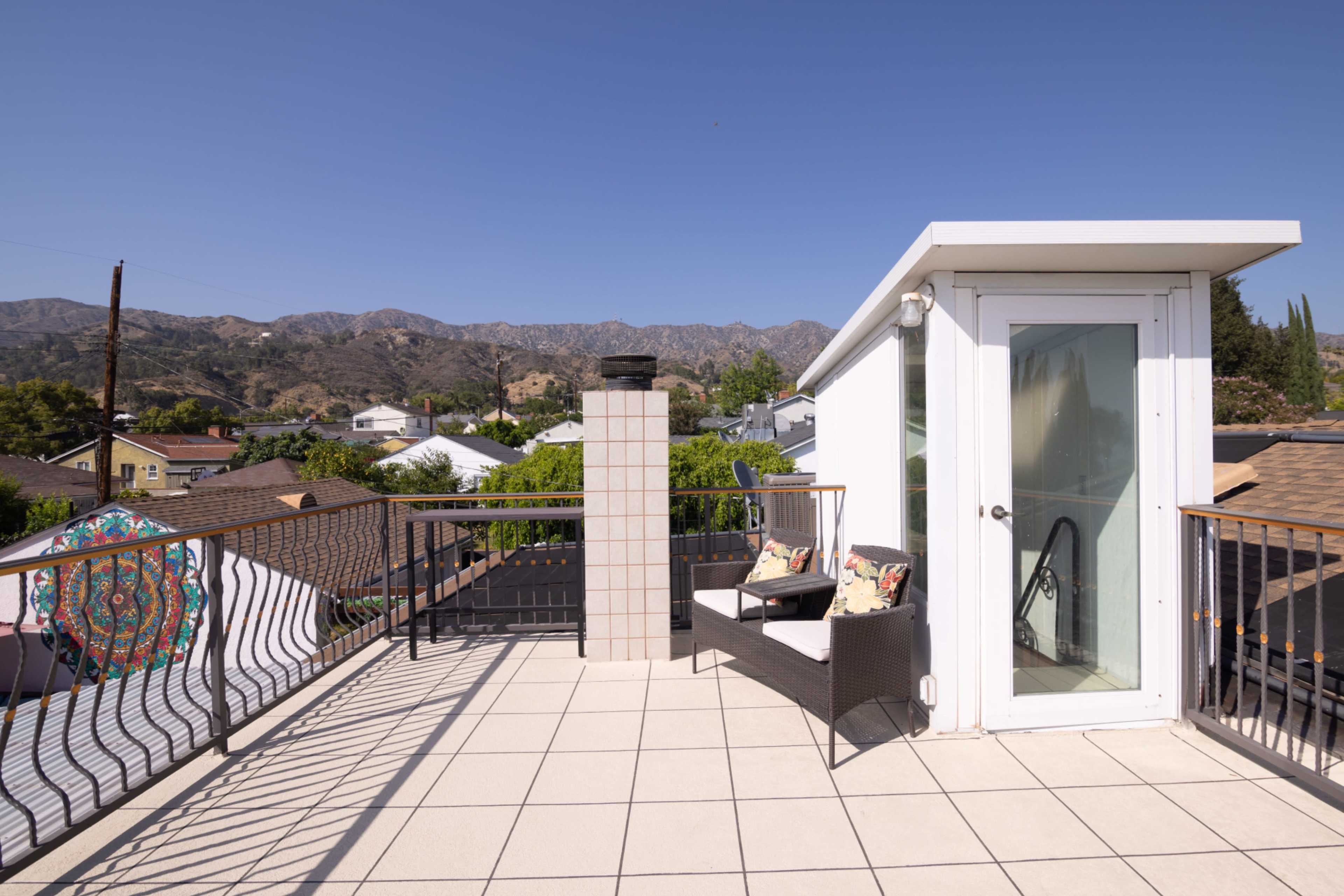 A rooftop terrace with a seating area, a chimney, and a mountainous backdrop under a clear blue sky.