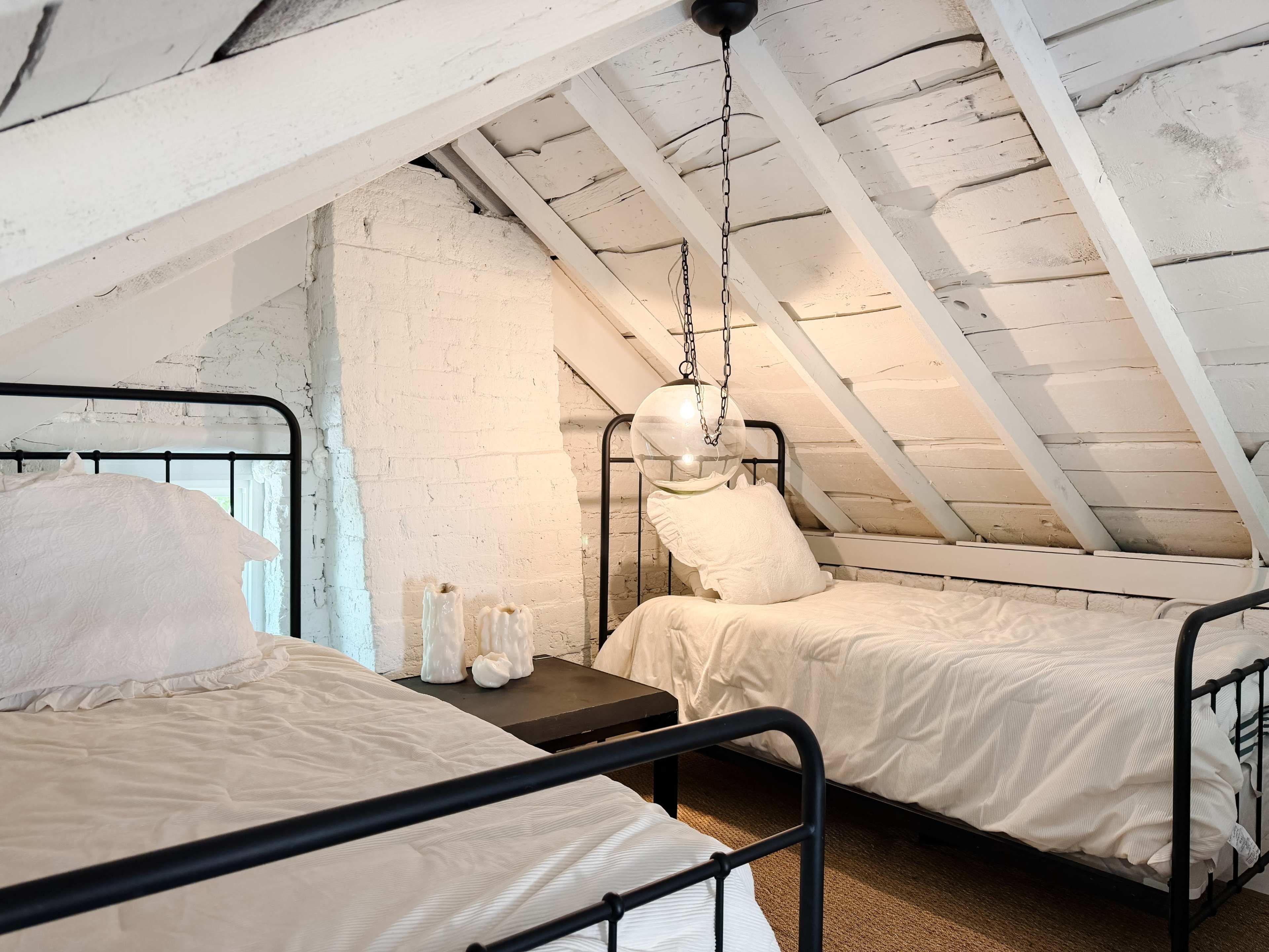 The image shows a cozy attic room featuring two iron bedframes with white bedding, a small black nightstand, and exposed wooden beams.